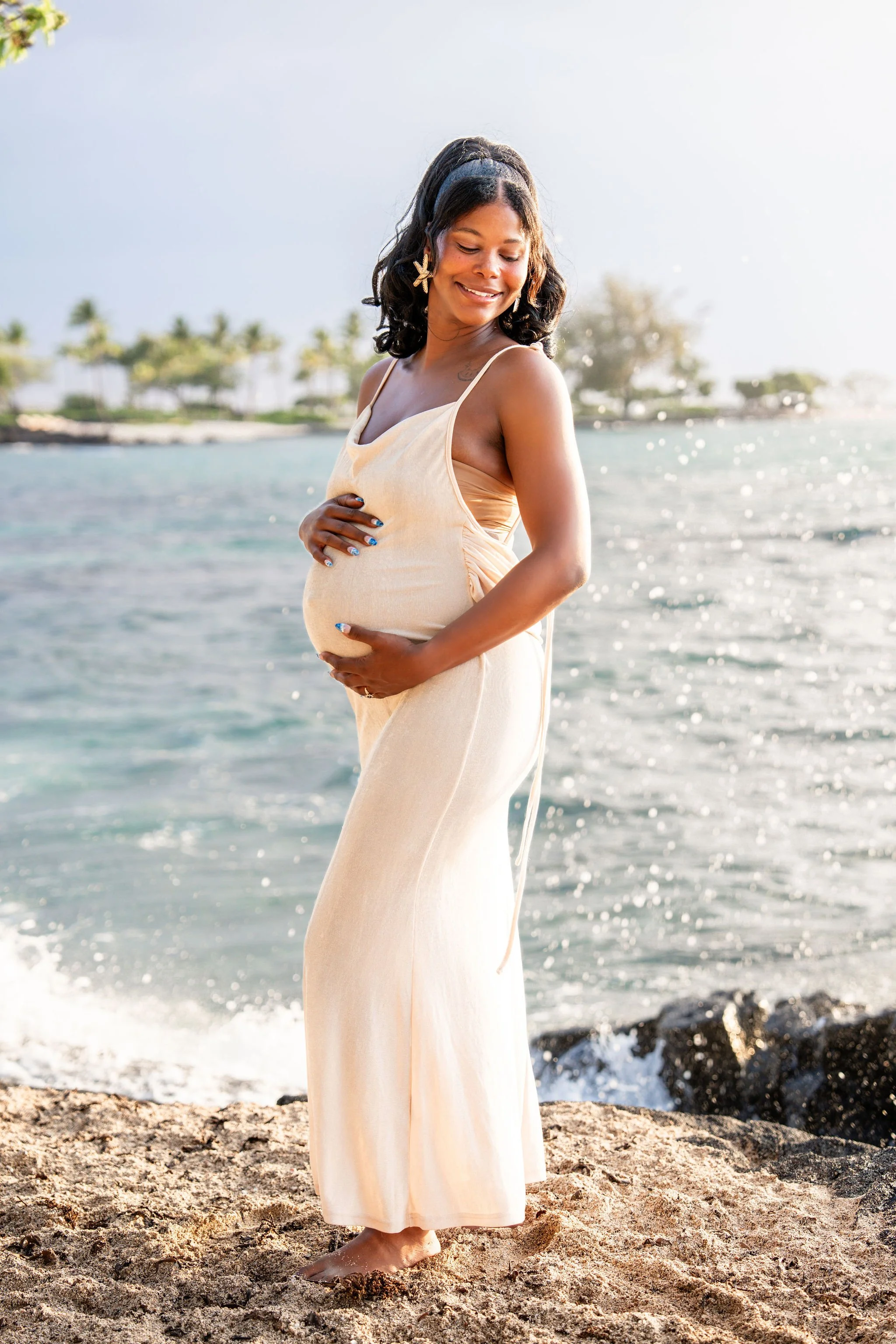A pregnant woman in a beige dress stands barefoot on a sandy beach near the water, cradling her baby bump with both hands and smiling.