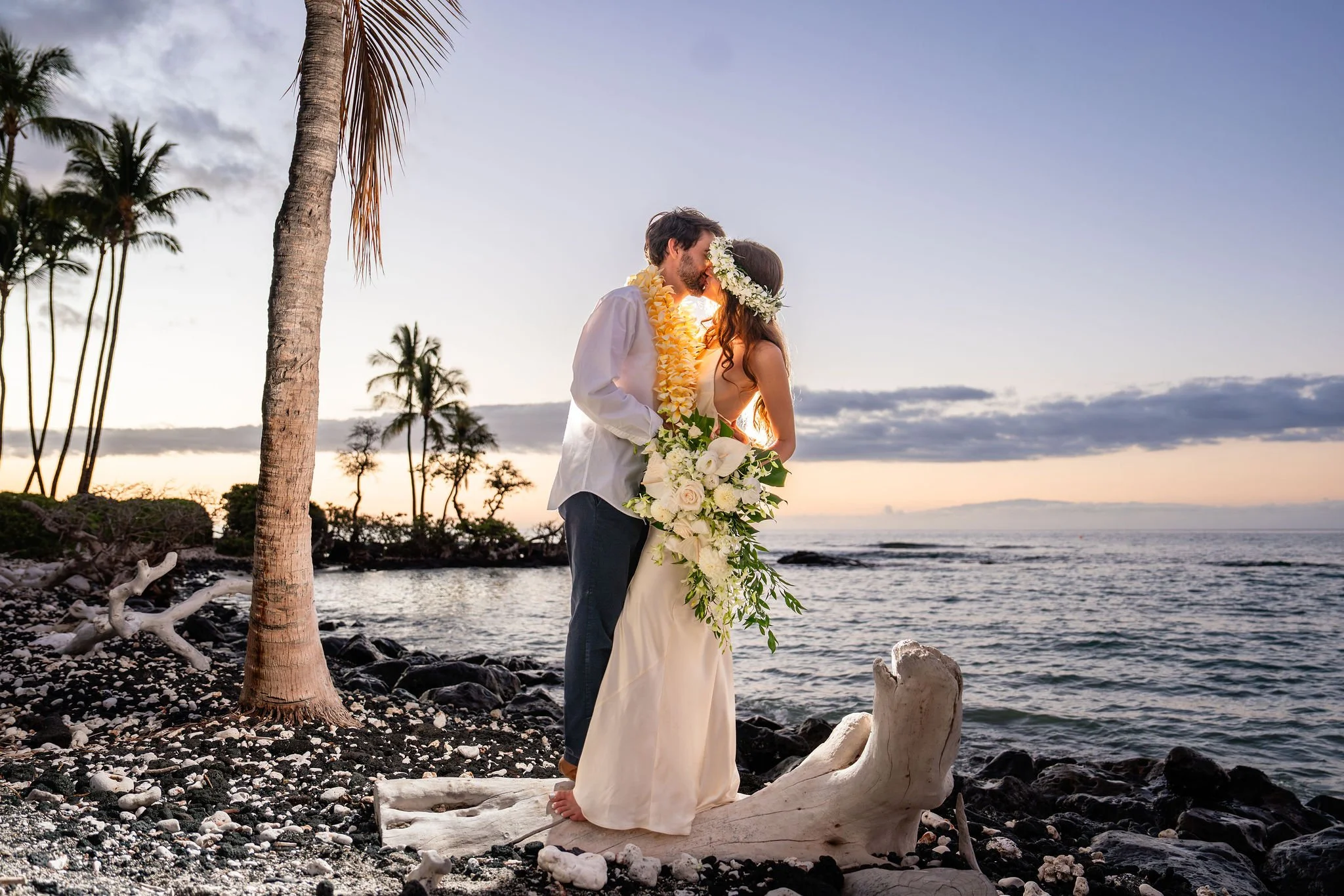 A couple in wedding attire sharing a kiss on a beach at sunset, with palm trees and a driftwood sculpture near the water.