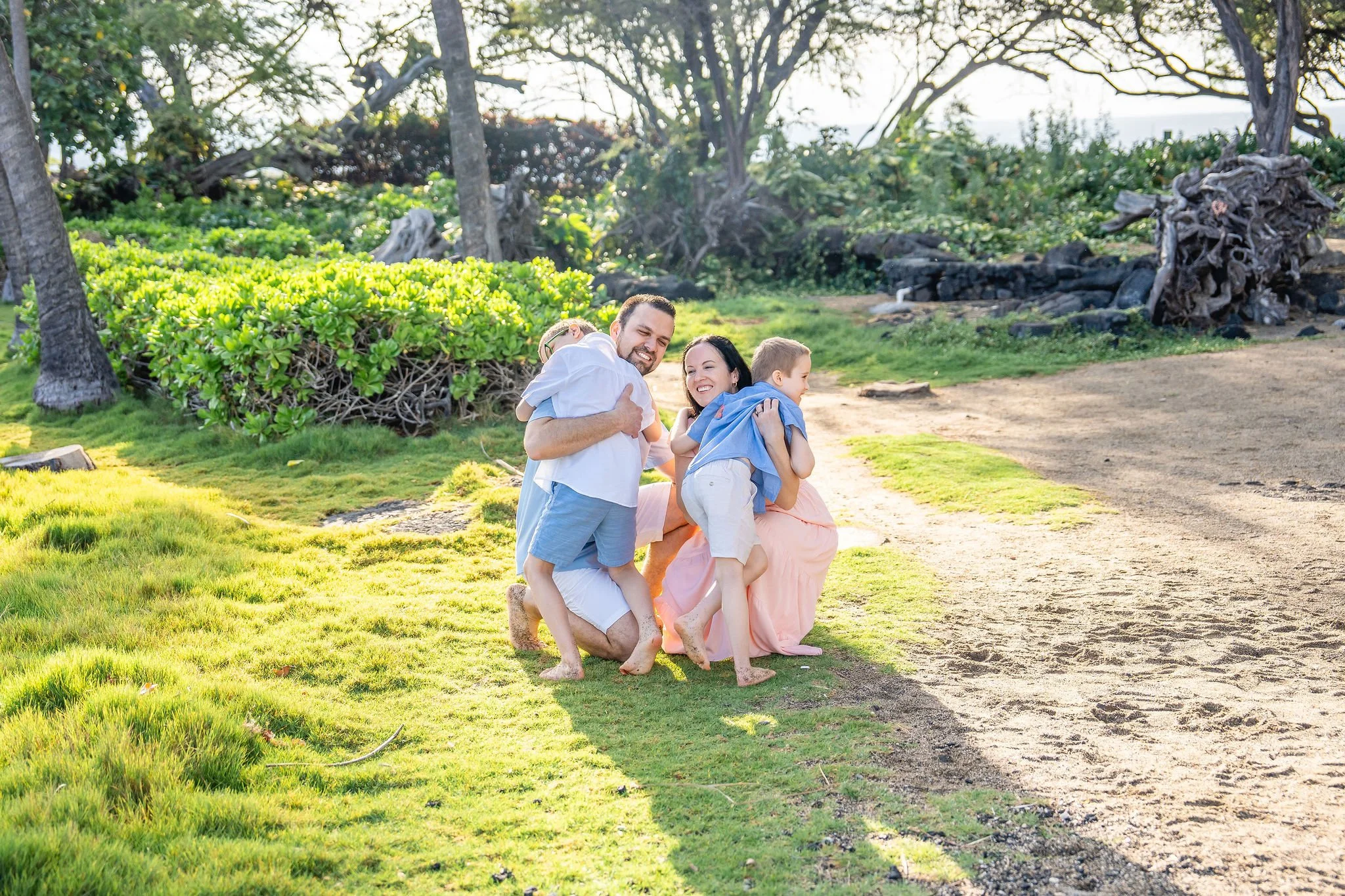 A family of four, including a man, woman, and two children, are playing together on a grassy area near trees in a park or backyard during sunset, smiling and hugging each other.