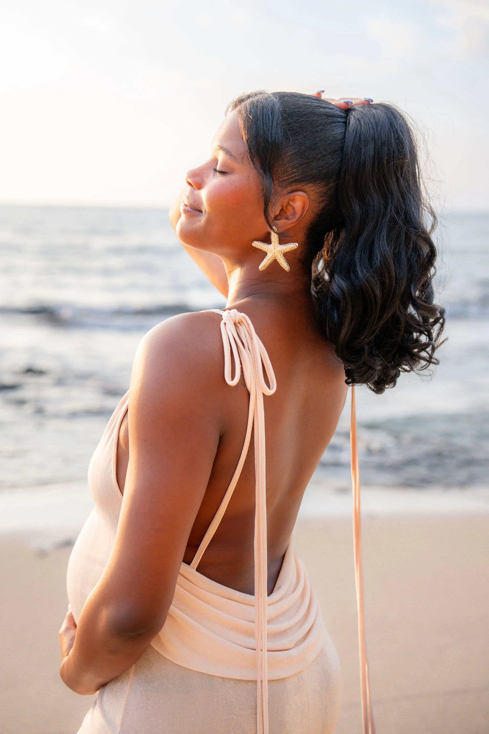 A woman with long dark hair styled in a high ponytail, wearing starfish earrings and a beige dress, stands on a beach with her eyes closed and a peaceful expression, holding her head with one hand, during sunset.