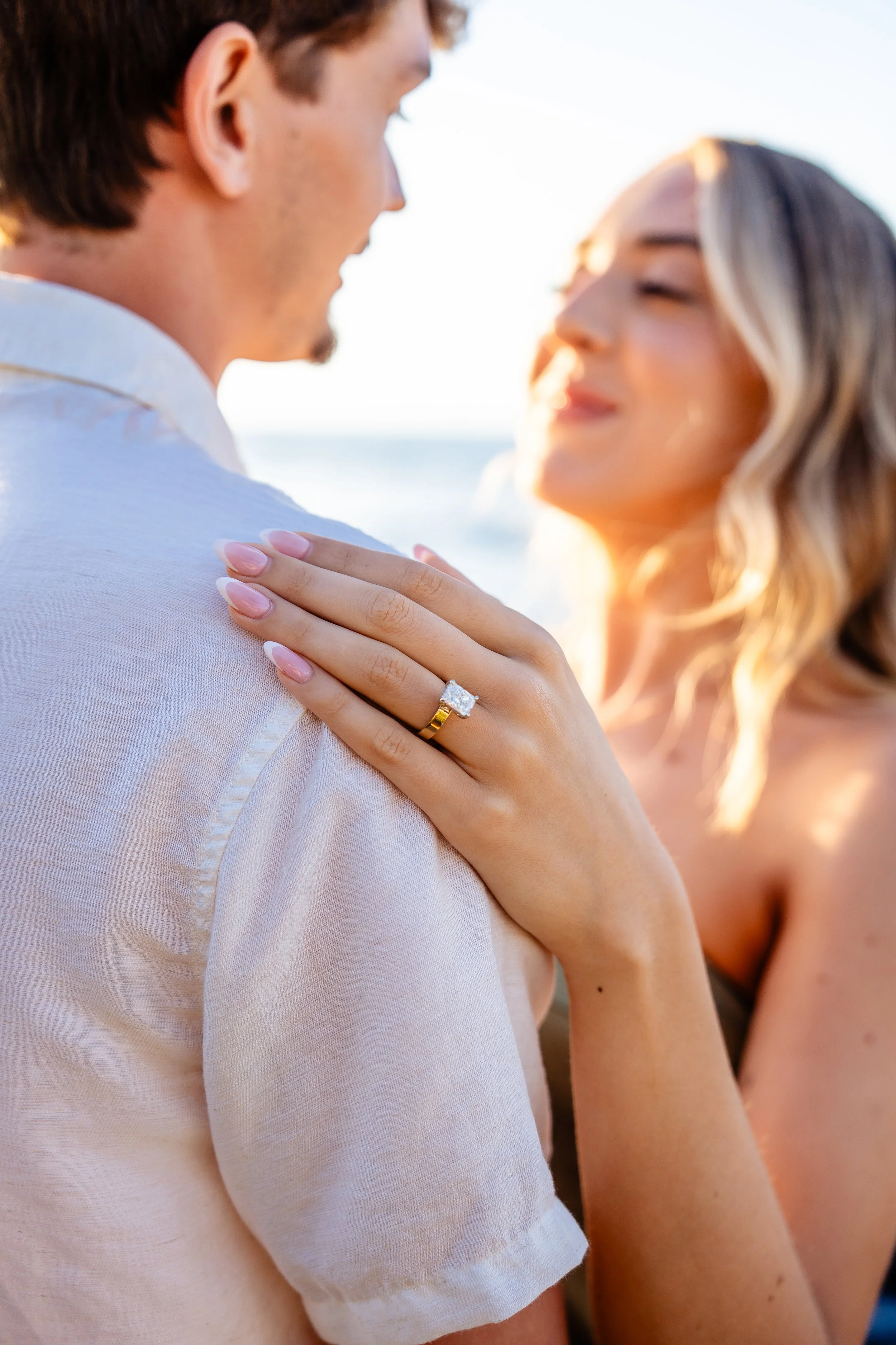 Close-up of a woman showing her left hand with an engagement ring resting on a man's shoulder, with the woman smiling and the man facing away, in an outdoor setting during sunset.
