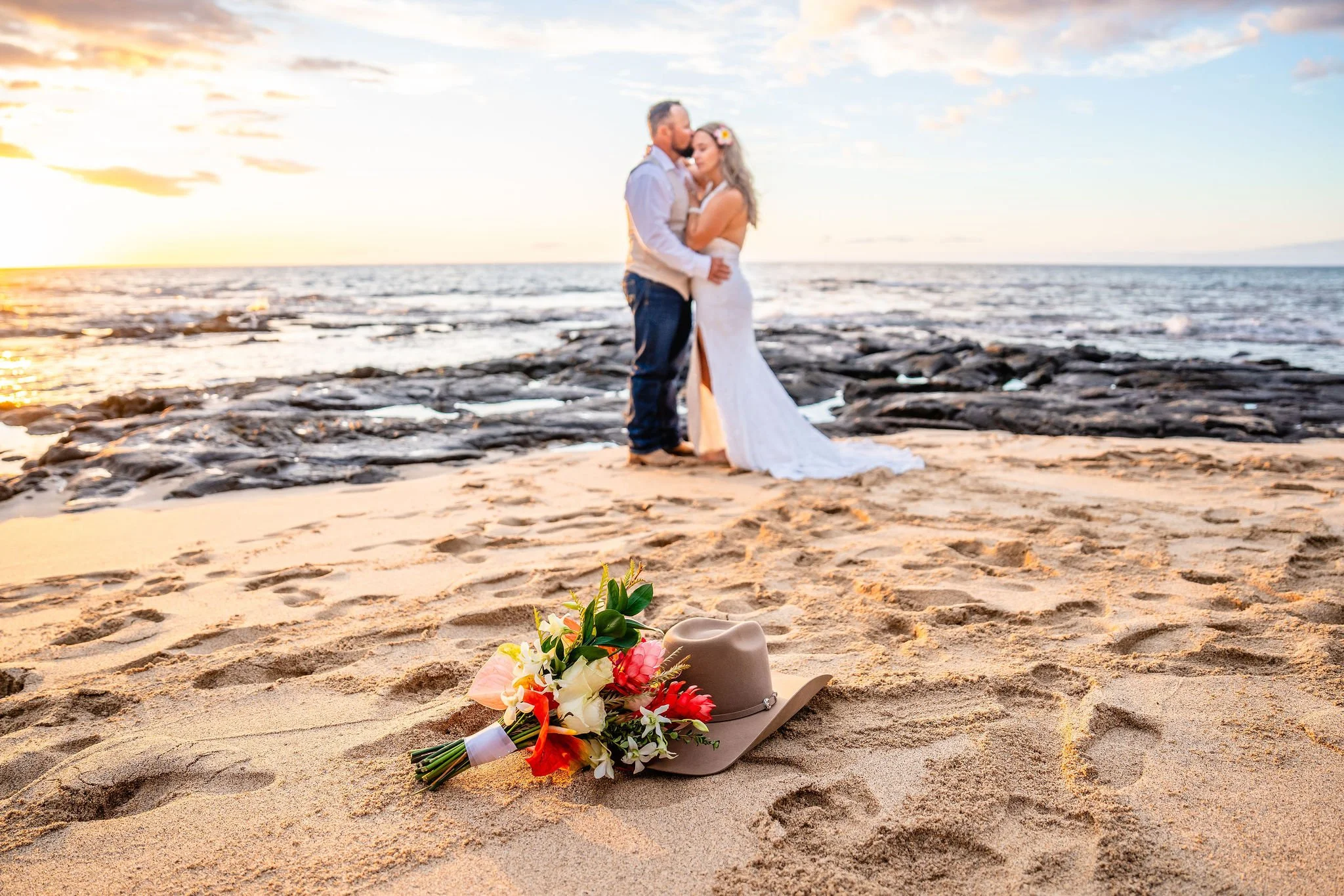 A wedding couple embracing on a beach at sunset, with a bouquet and a hat with flowers placed on the sand in the foreground.