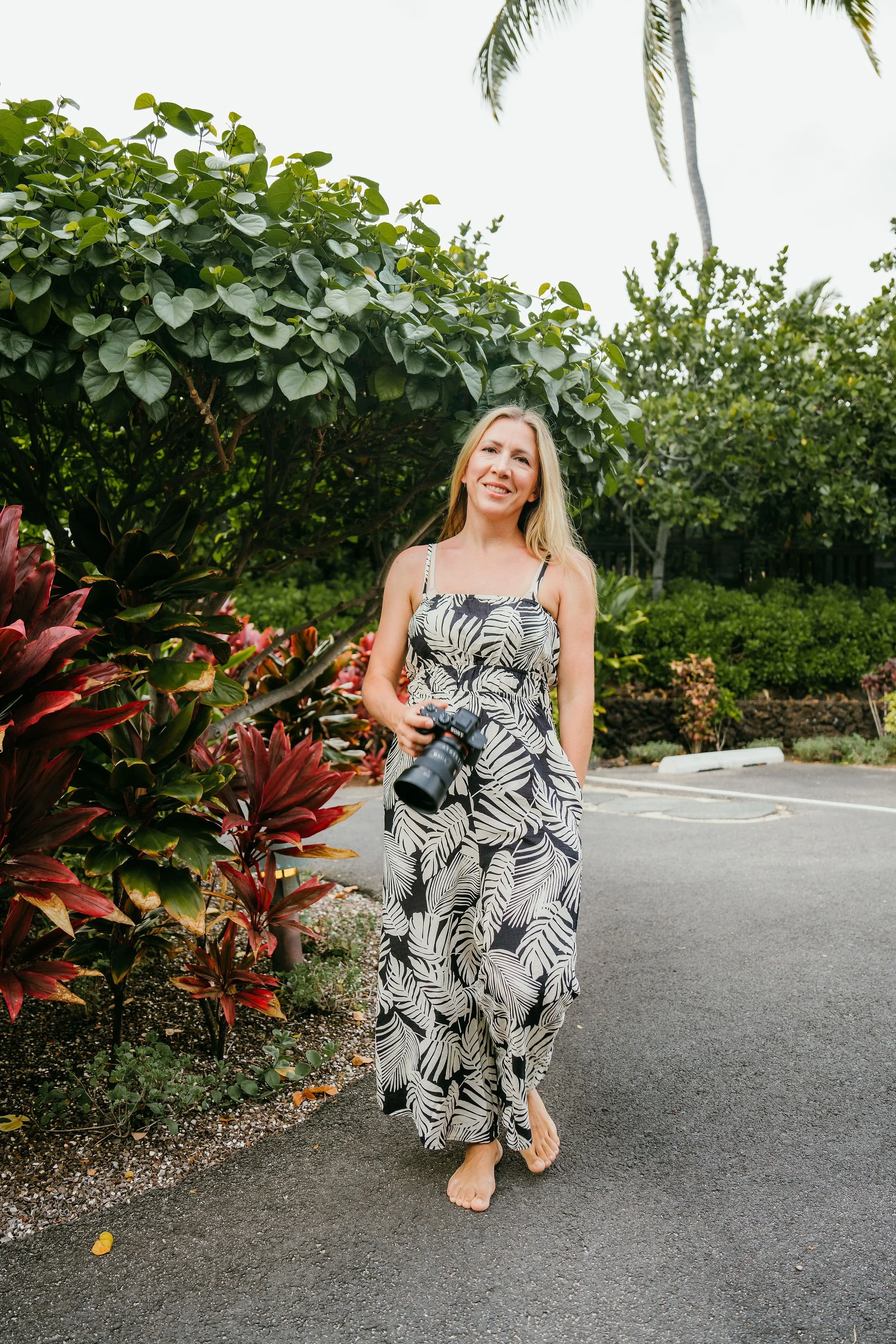 A woman with long blonde hair standing outdoors barefoot next to a garden bed with red and green leafy plants, holding a camera, wearing a black and white tropical print dress, smiling with a background of green trees and a palm tree.