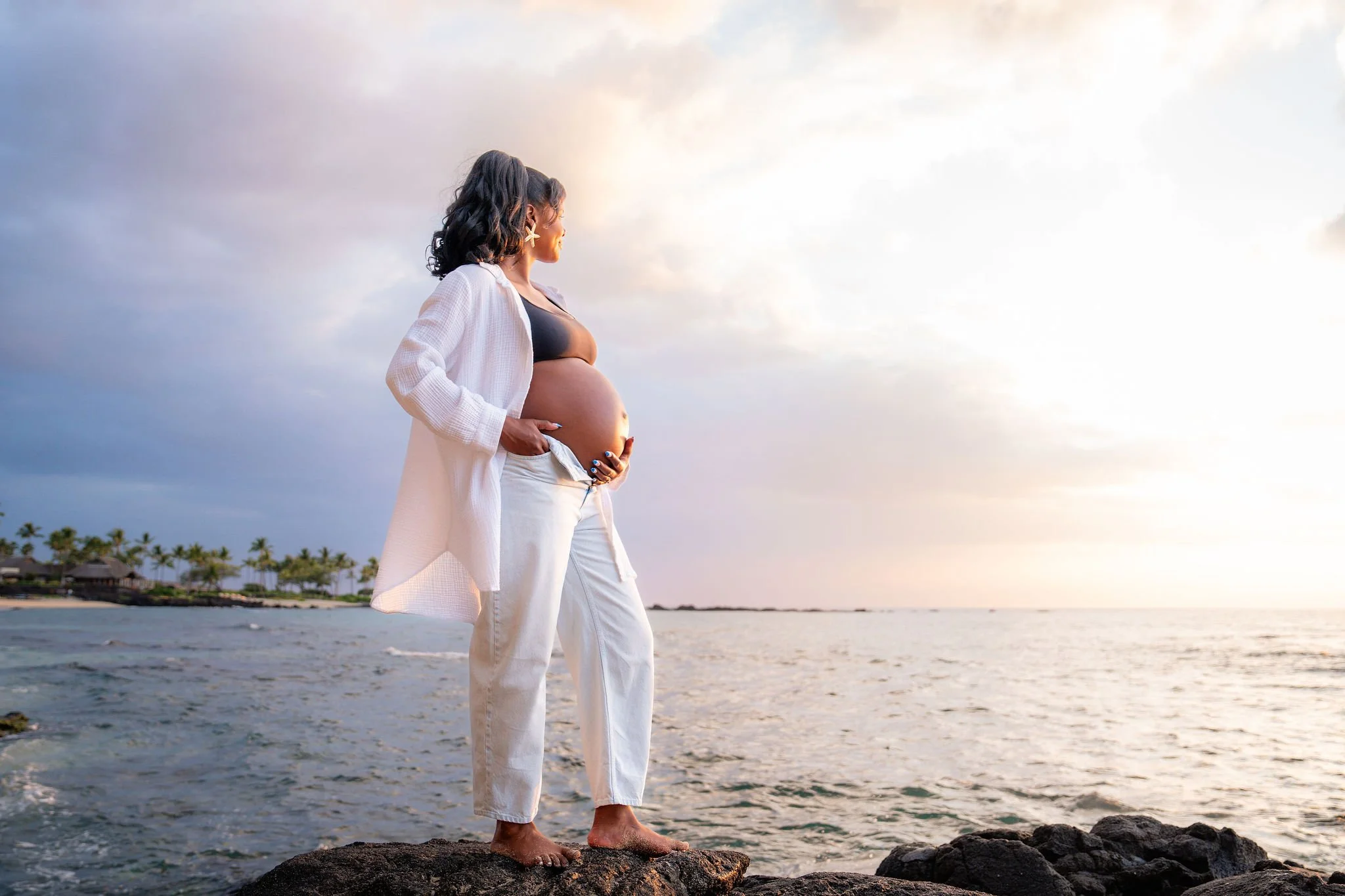 A pregnant woman with dark hair, wearing a black top and white pants, stands barefoot on rocks by the ocean at sunset, looking towards the horizon with her hands on her belly.