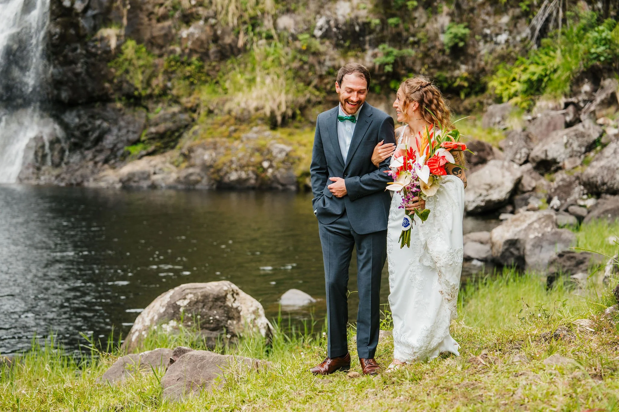 A newlywed couple dressed in wedding attire stands near a river or pond with a waterfall in the background. The groom is in a dark suit with a green bow tie, and the bride is holding a large bouquet of colorful flowers. They are smiling and looking at each other, surrounded by rocks and green grass.