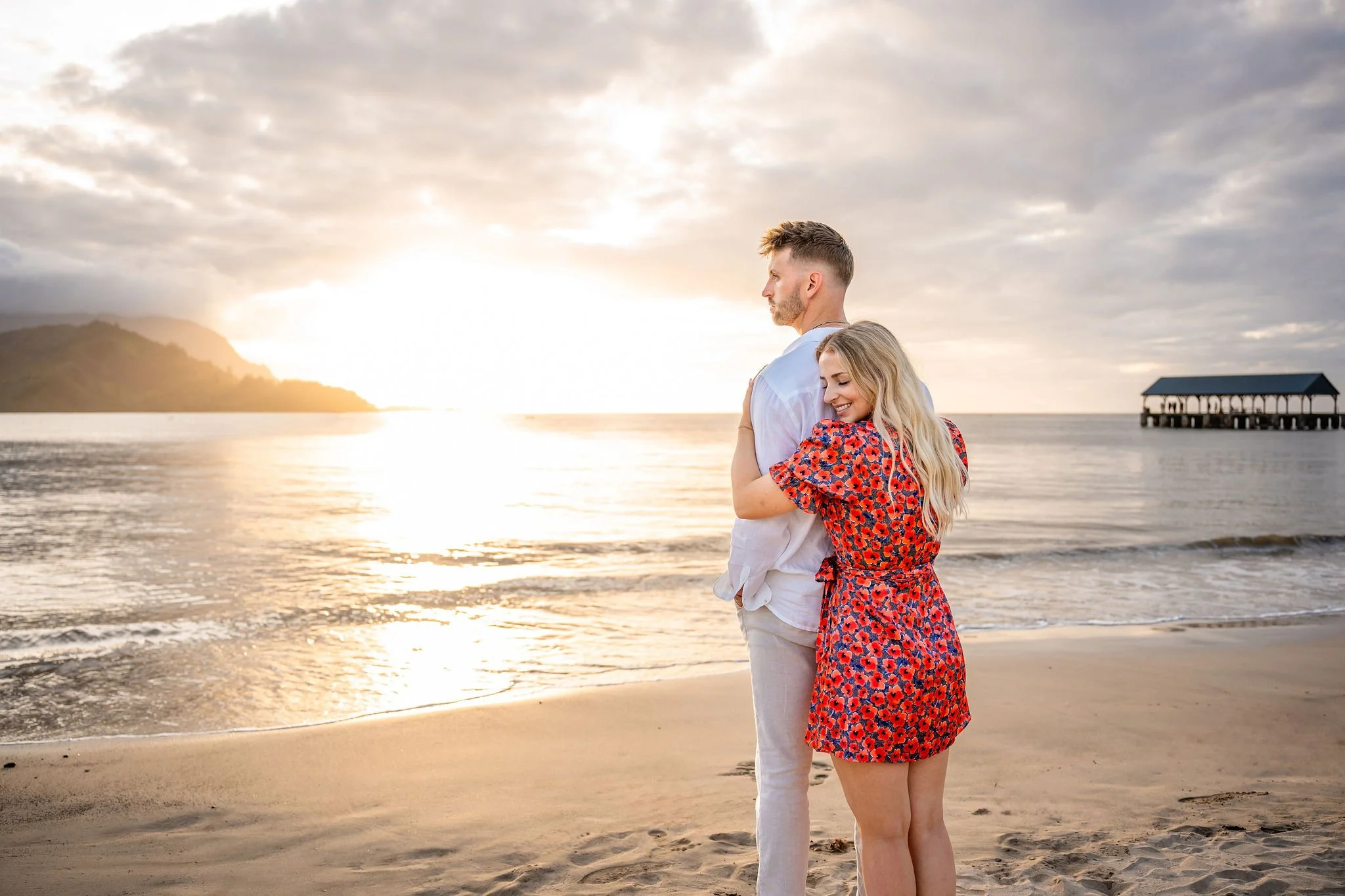 A man and woman standing on the beach at sunset, embracing each other, with waves and a pier in the background.