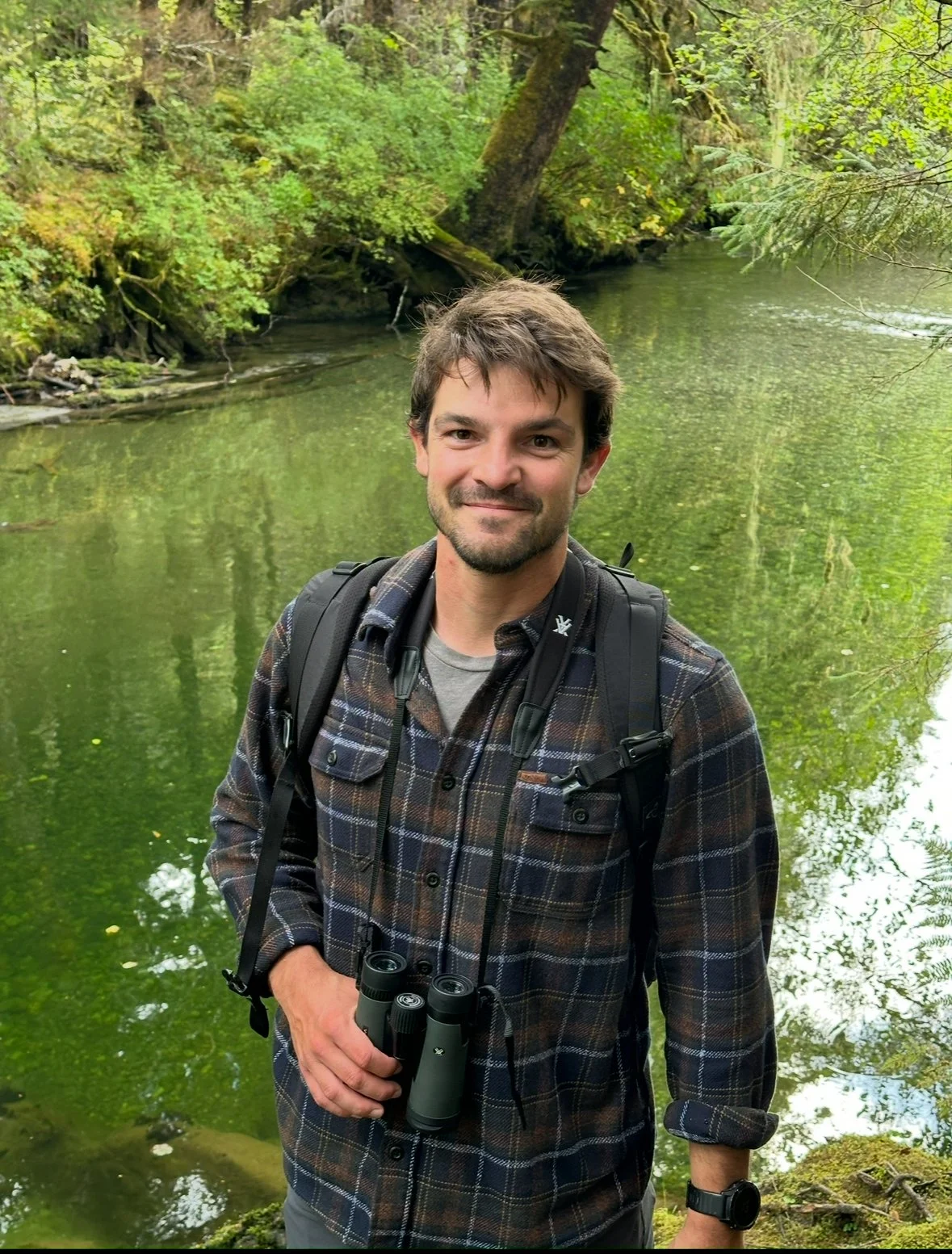 A man with short brown hair and a beard smiling, standing near a green river in a wooded area, holding binoculars. He is wearing a plaid shirt, a backpack, and a watch.