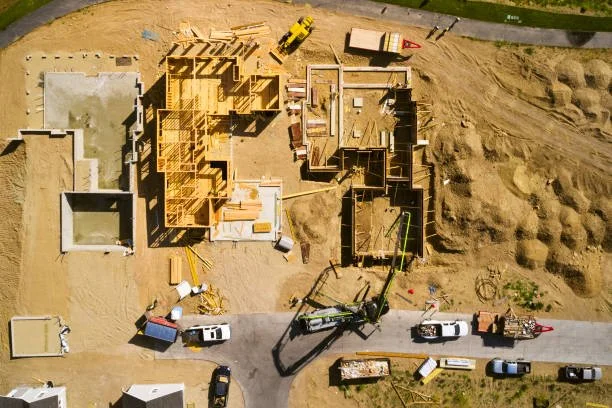 Aerial view of a construction site showing multiple wooden structures in different stages of building, construction vehicles, and equipment, with a parking lot and dirt areas surrounding the site.