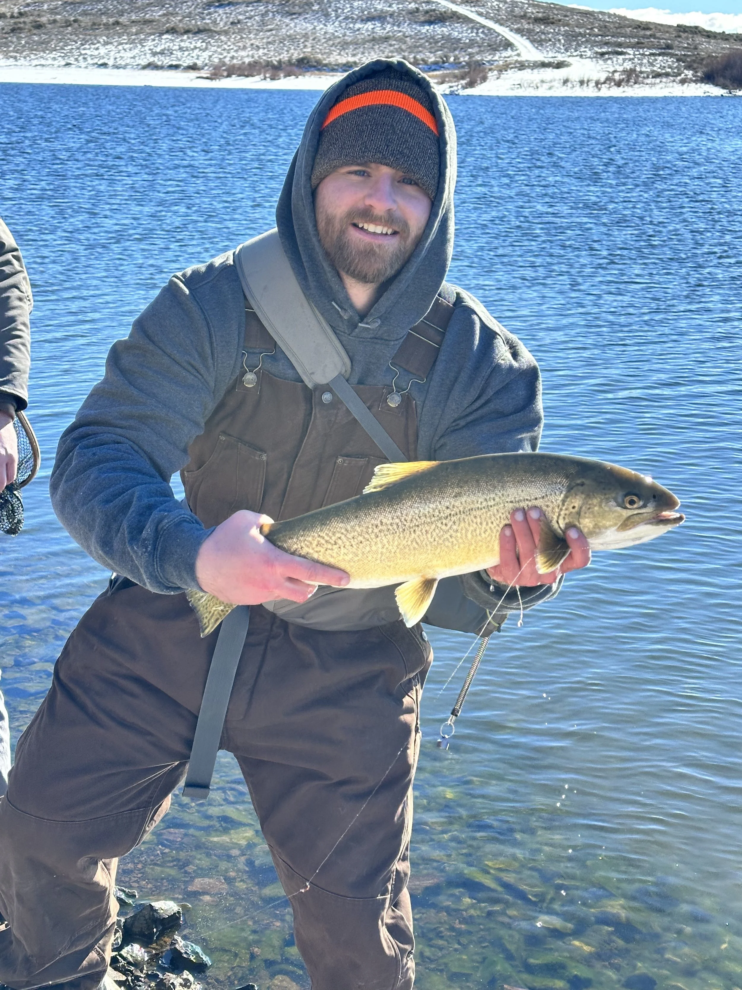 A man wearing layered outdoor clothing, including a beanie and hoodie, is smiling while holding a large fish with a yellowish body. He is standing in shallow water near rocky terrain, with a large blue body of water and snow-covered hills in the background.
