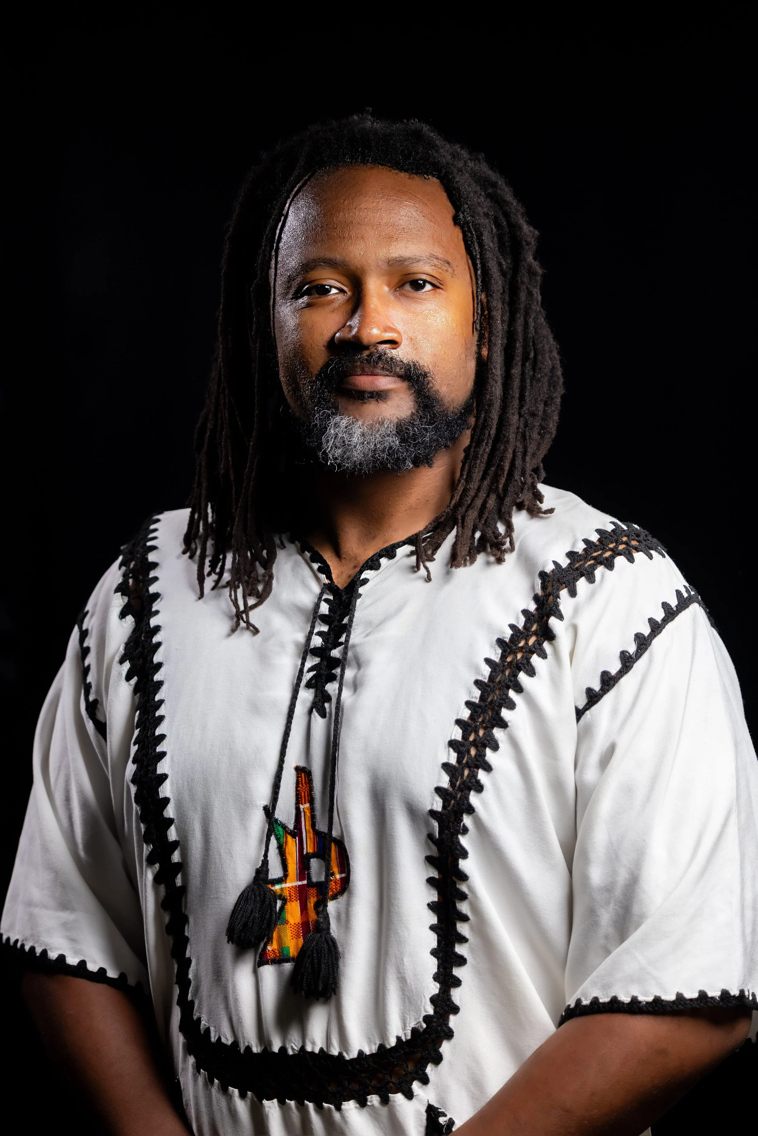 A man with dreadlocks, a beard with gray, and medium-dark skin, wearing a traditional embroidered white shirt with black stitching, stands against a black background.