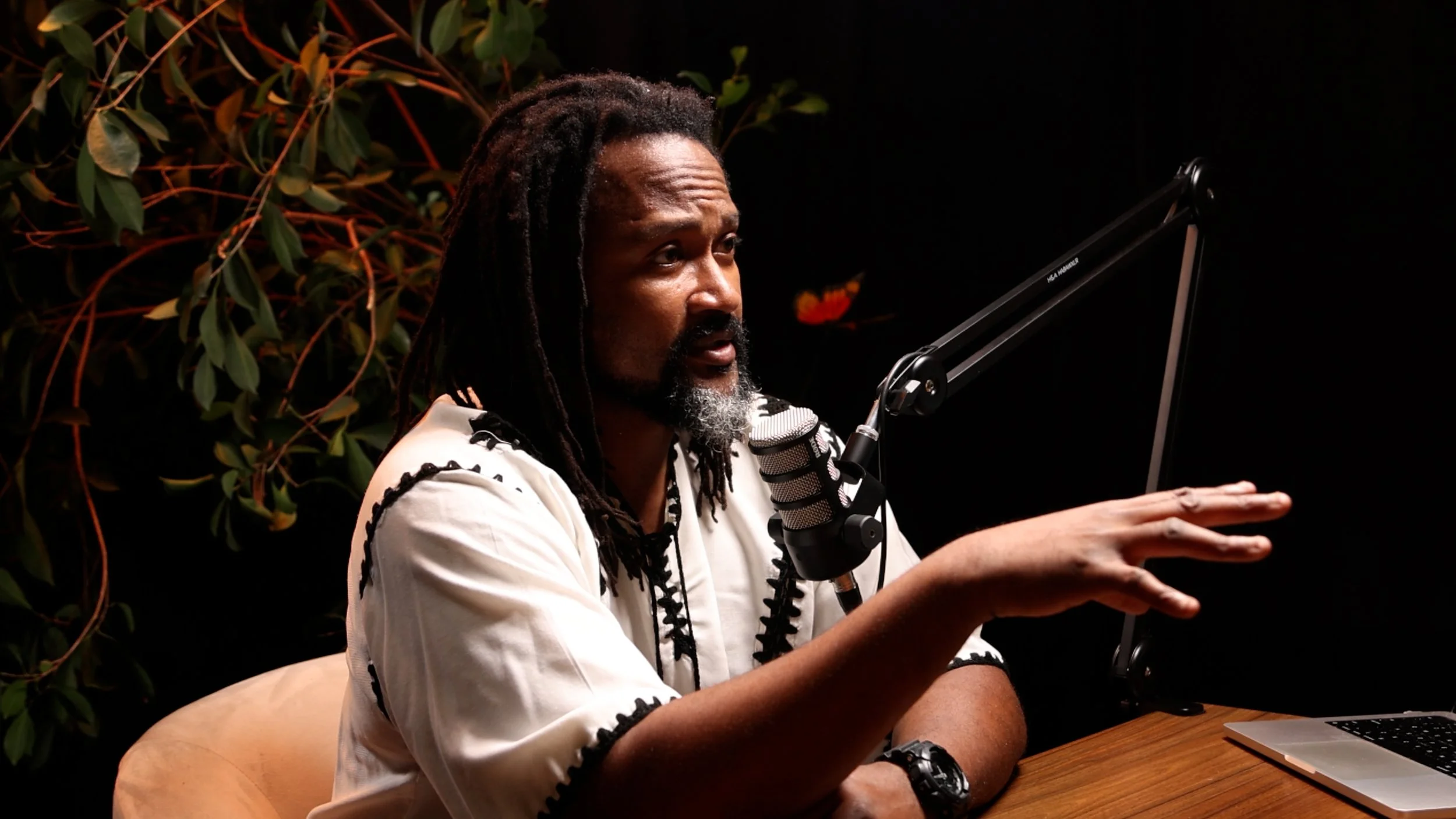 A man with dreadlocks and a beard speaks into a microphone while gesturing with his hand, sitting at a wooden table with a laptop, against a black background with green leaves.