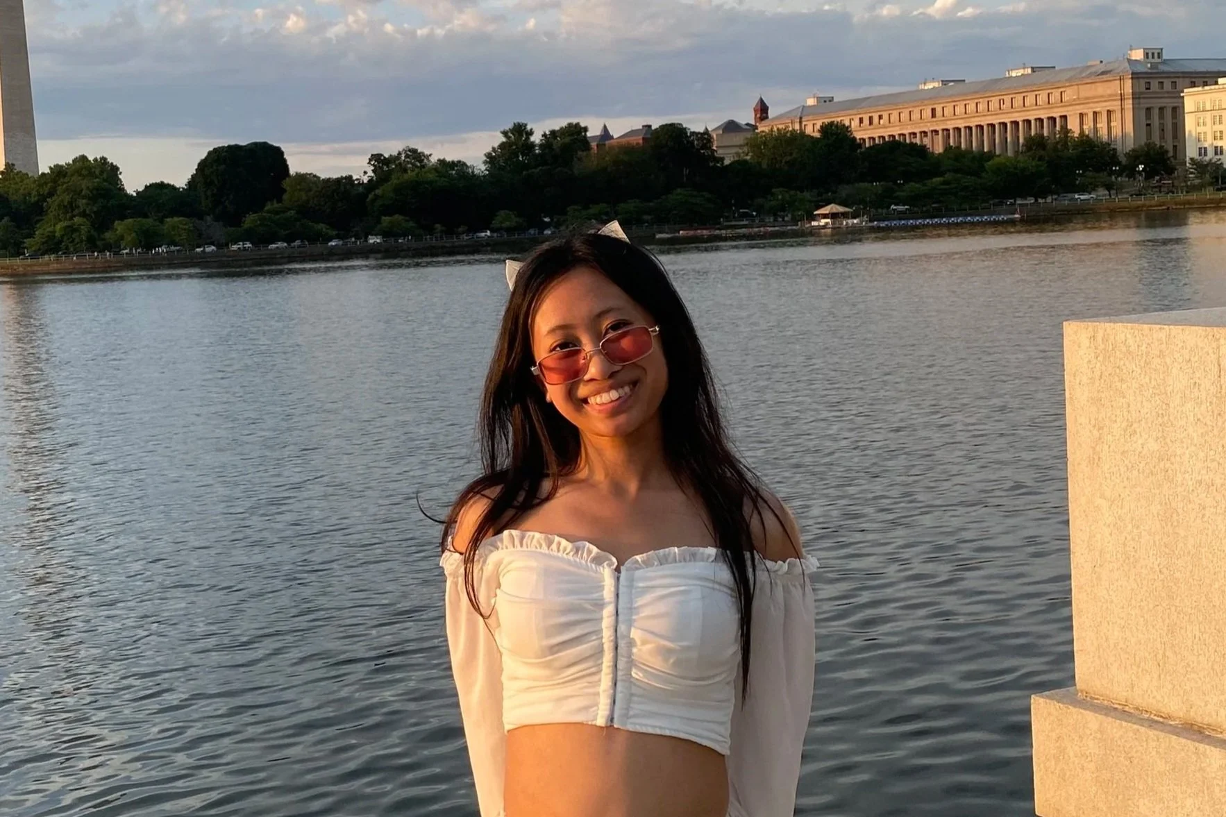 Pictured is Quenzy with long dark hair wearing pink-tinted sunglasses and a white off-the-shoulder crop top, smiling by a body of water at sunset with trees and large buildings in the background.