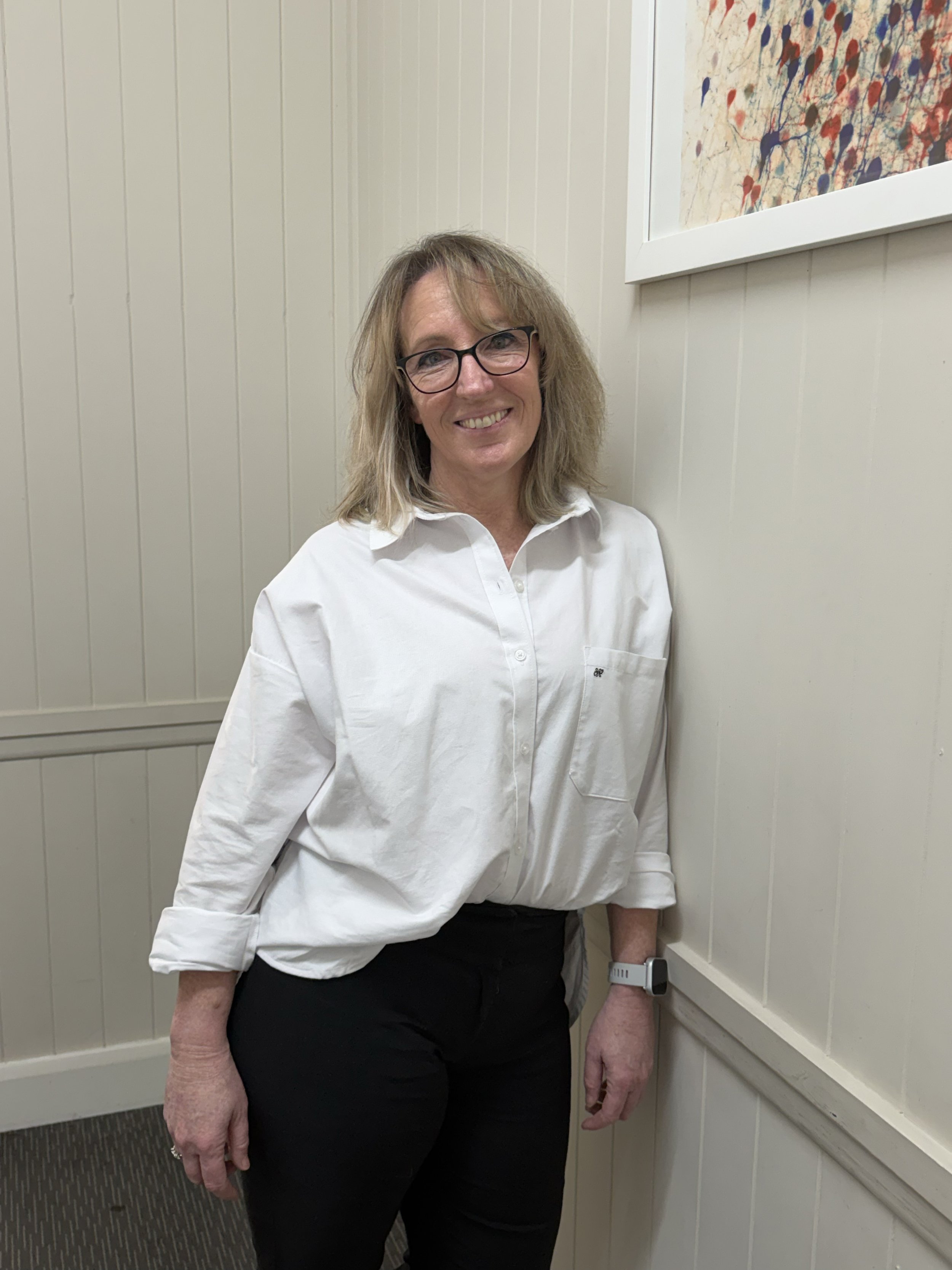 A woman with shoulder-length blonde hair, wearing glasses, a white shirt, and black pants, standing against cream-colored, vertically paneled walls with a framed abstract painting above her right shoulder.