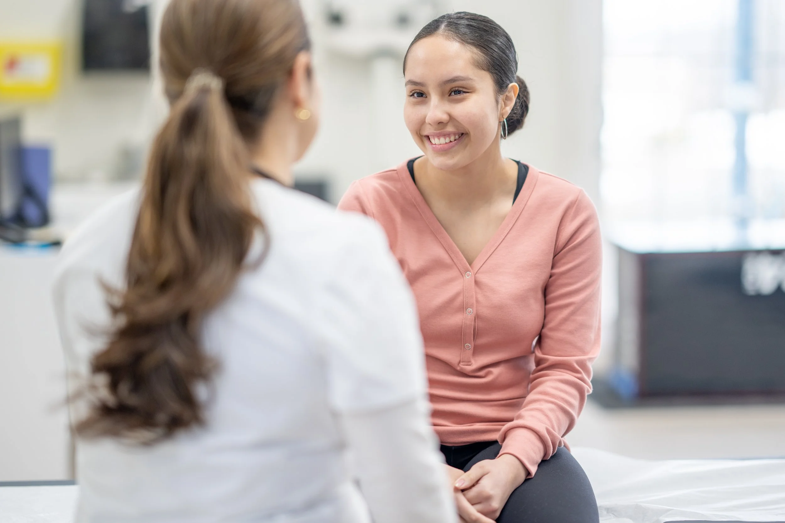 A female doctor having a conversation with a female teenager in a room, one facing the camera with a smiling expression, the other with her back to the camera, slightly out of focus.