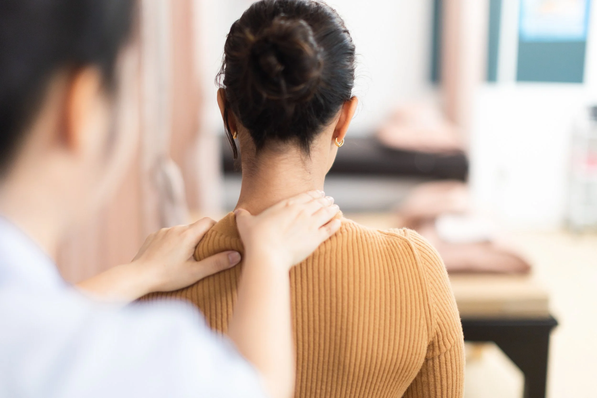 A woman with dark hair in a bun receiving a shoulder treatment massage from a physiotherapist in a bright, indoor setting.