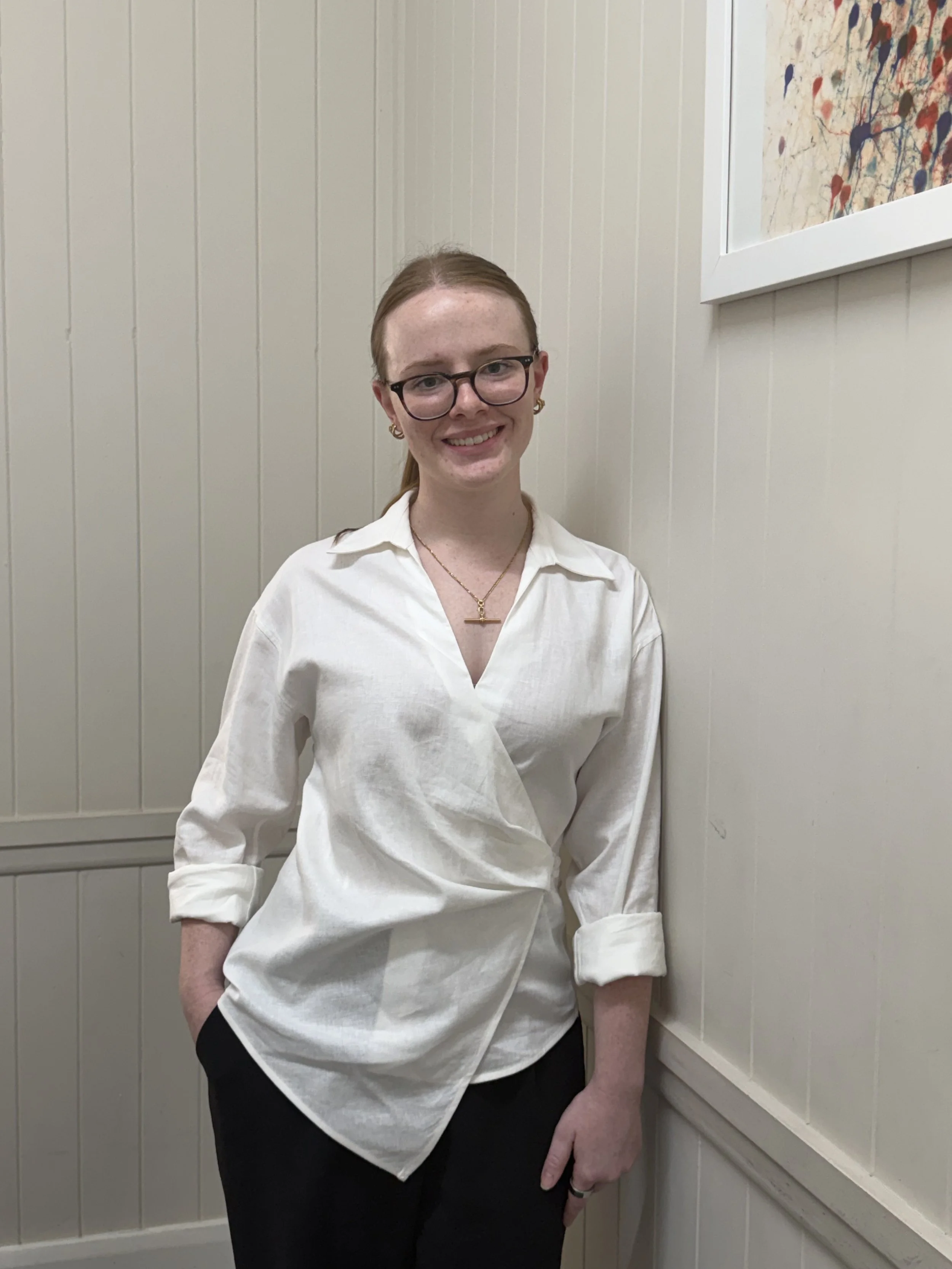 A woman with auburn hair pulled back, wearing glasses, a white shirt with rolled-up sleeves, and gold jewellery, standing against a beige paneled wall with a colorful abstract painting in a white frame.