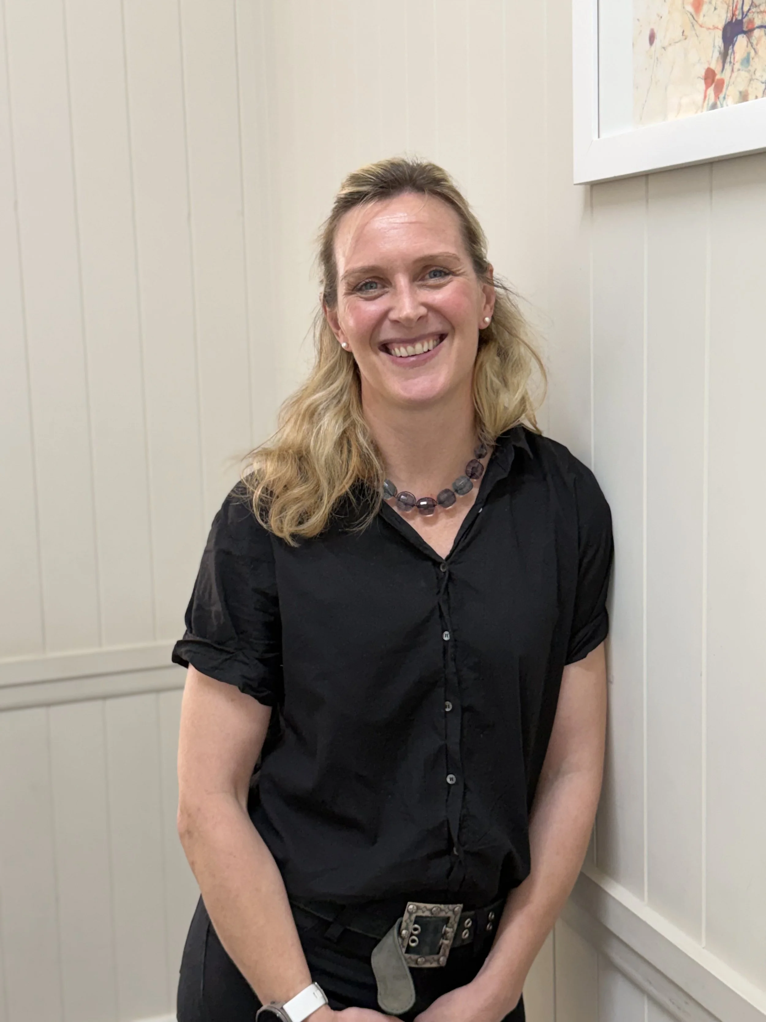 A woman with blonde hair smiling, standing indoors against a beige wall with a colorful abstract painting on the wall to her right. She is wearing a black shirt, a beaded necklace, and a white watch.