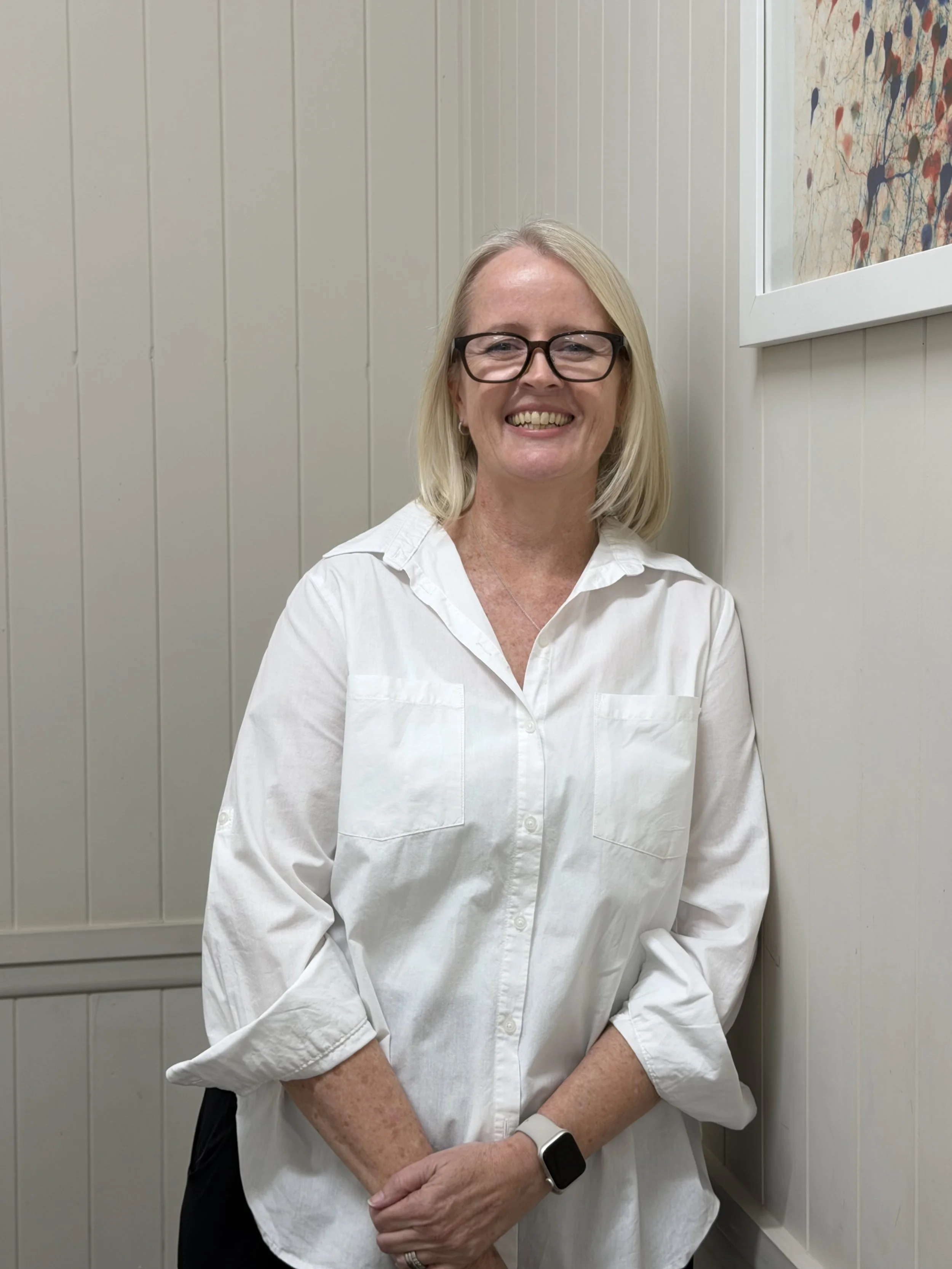 A smiling woman with blonde hair, glasses, and a white button-up shirt standing against a white paneled wall with a colorful abstract painting on the right side of the image.