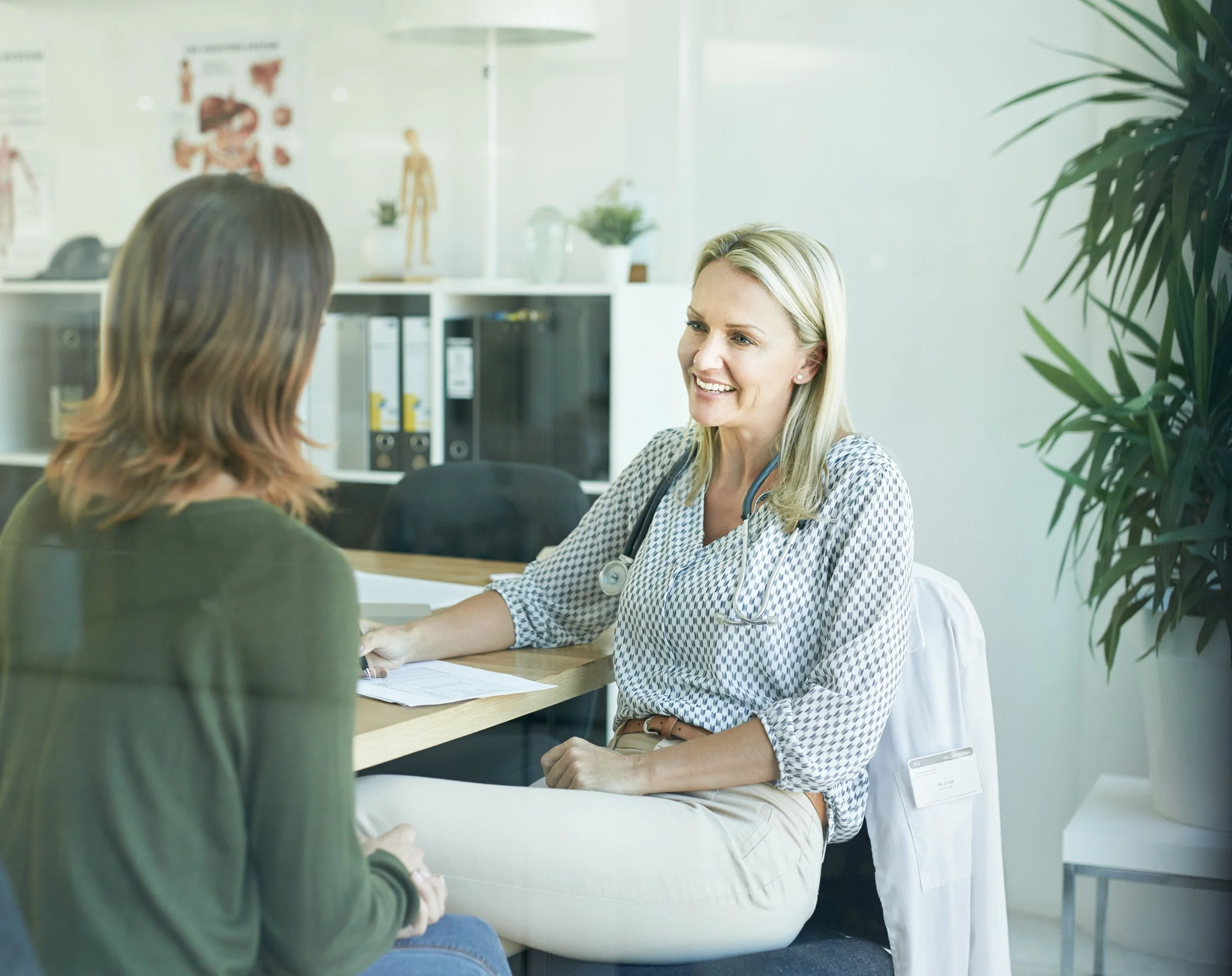 A healthcare professional, smiling and engaging in a consultation with a patient in a medical office. The healthcare provider is wearing a stethoscope around her neck and has a name badge, while the patient is sitting across from her with her back to the camera. The office has medical posters, bookshelves with binders, and a large green plant in the foreground.