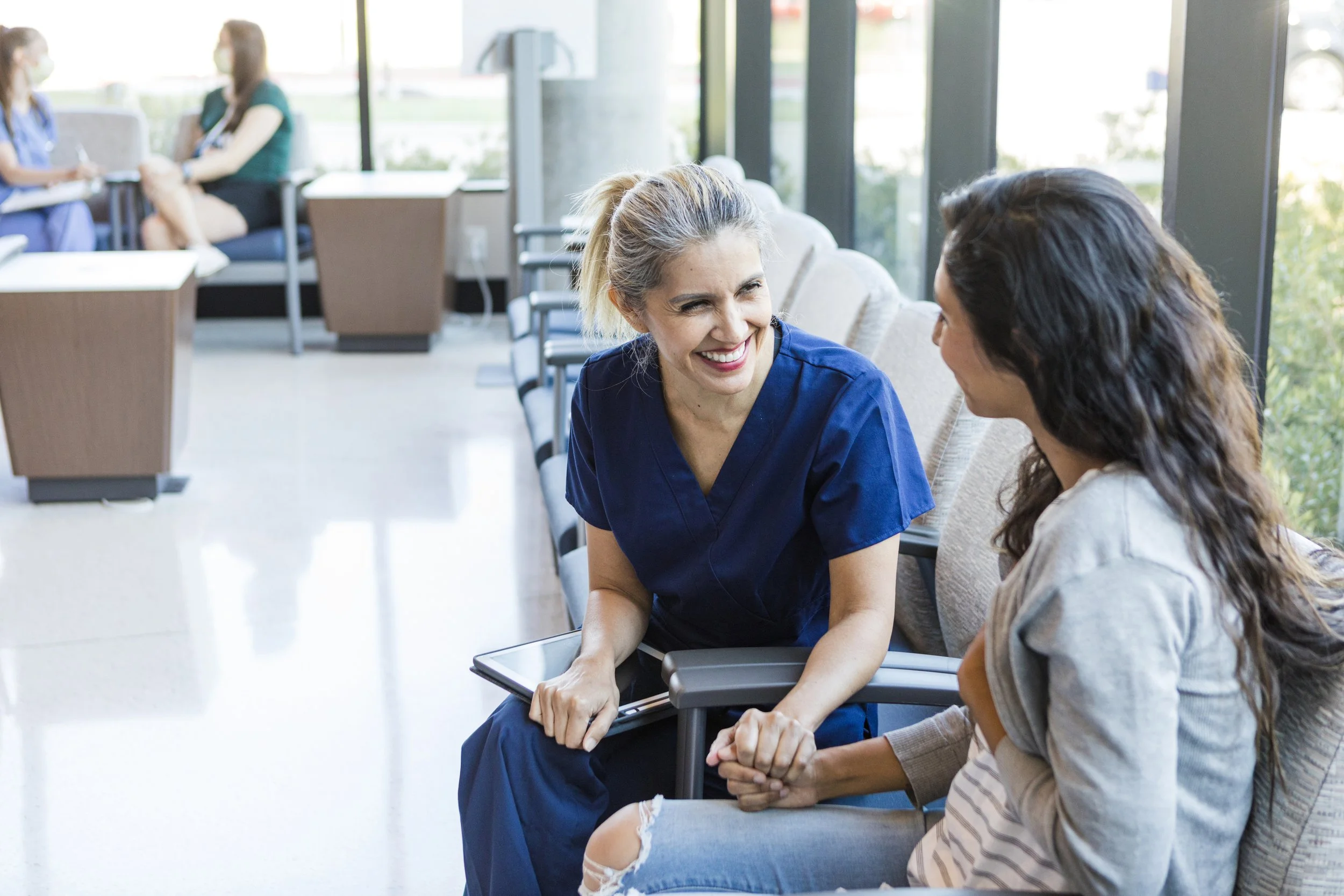 A smiling nurse in blue scrubs and a woman with long dark hair engaged in conversation, sitting on chairs in a medical waiting area with large windows, during daytime.