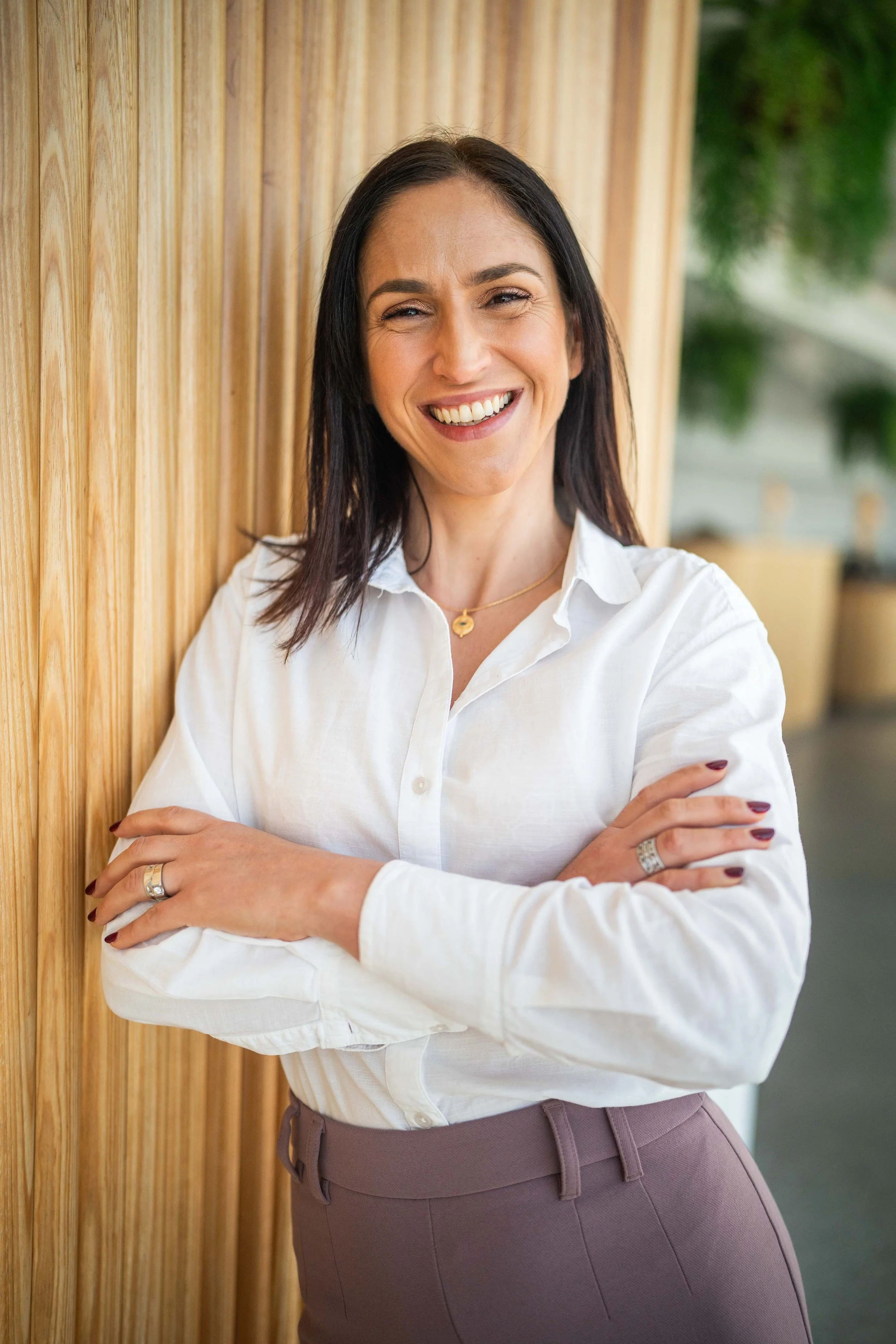 A woman with long dark hair, smiling, wearing a white shirt, sunlight marks on her face, in front of a wooden wall with green blurred background.