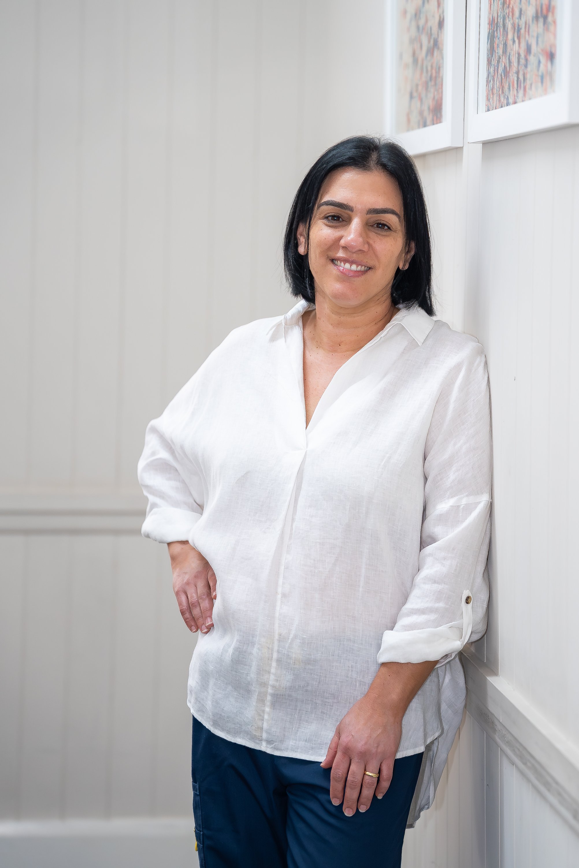 A woman with short black hair, wearing a white button-up shirt and dark pants, standing against a white wall with framed art. She is smiling, with her right hand on her hip and her left hand resting on her thigh.
