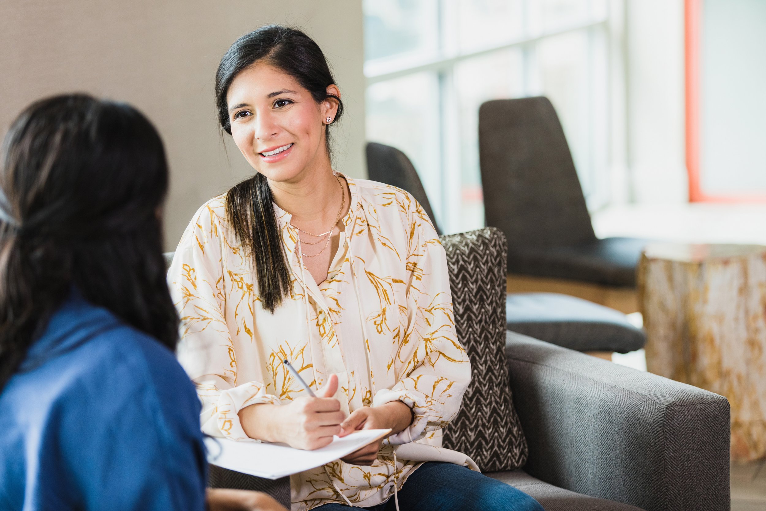 A woman with dark hair and wearing a light-colored patterned blouse is smiling and talking to another woman with dark hair wearing a blue top, seated in a room with large windows and chairs.