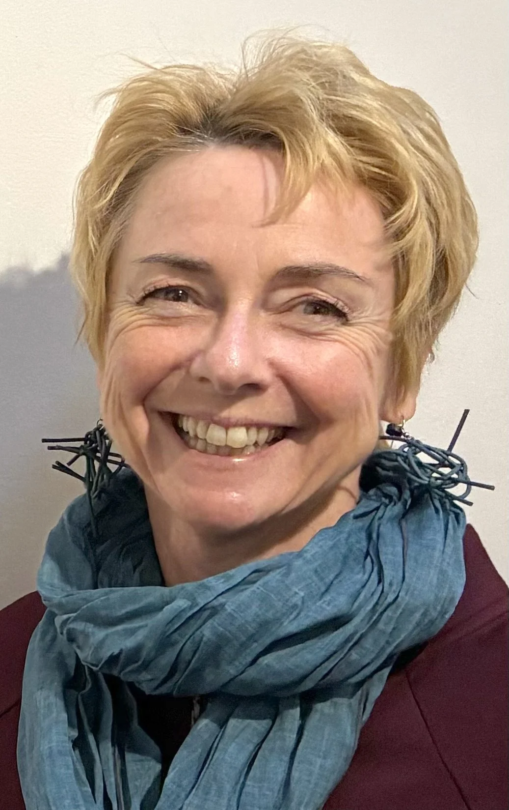 A smiling woman with short blonde hair, wearing a maroon top and a blue scarf around her neck, standing against a plain light-colored wall.