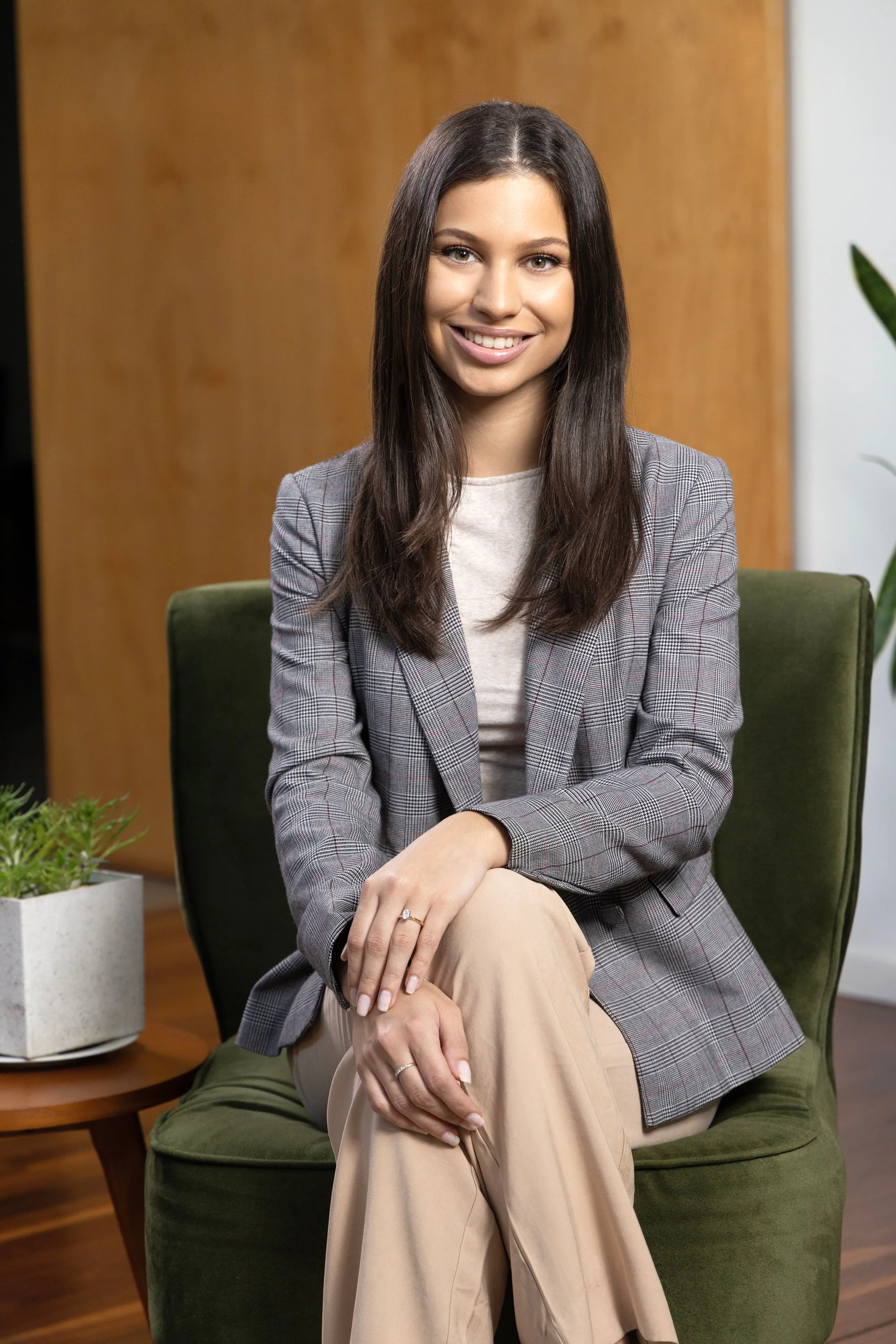 A young woman with long dark hair, wearing a gray plaid blazer and beige pants, smiling and sitting cross-legged on a green armchair in a modern office setting with a wooden wall and potted plant in the background.