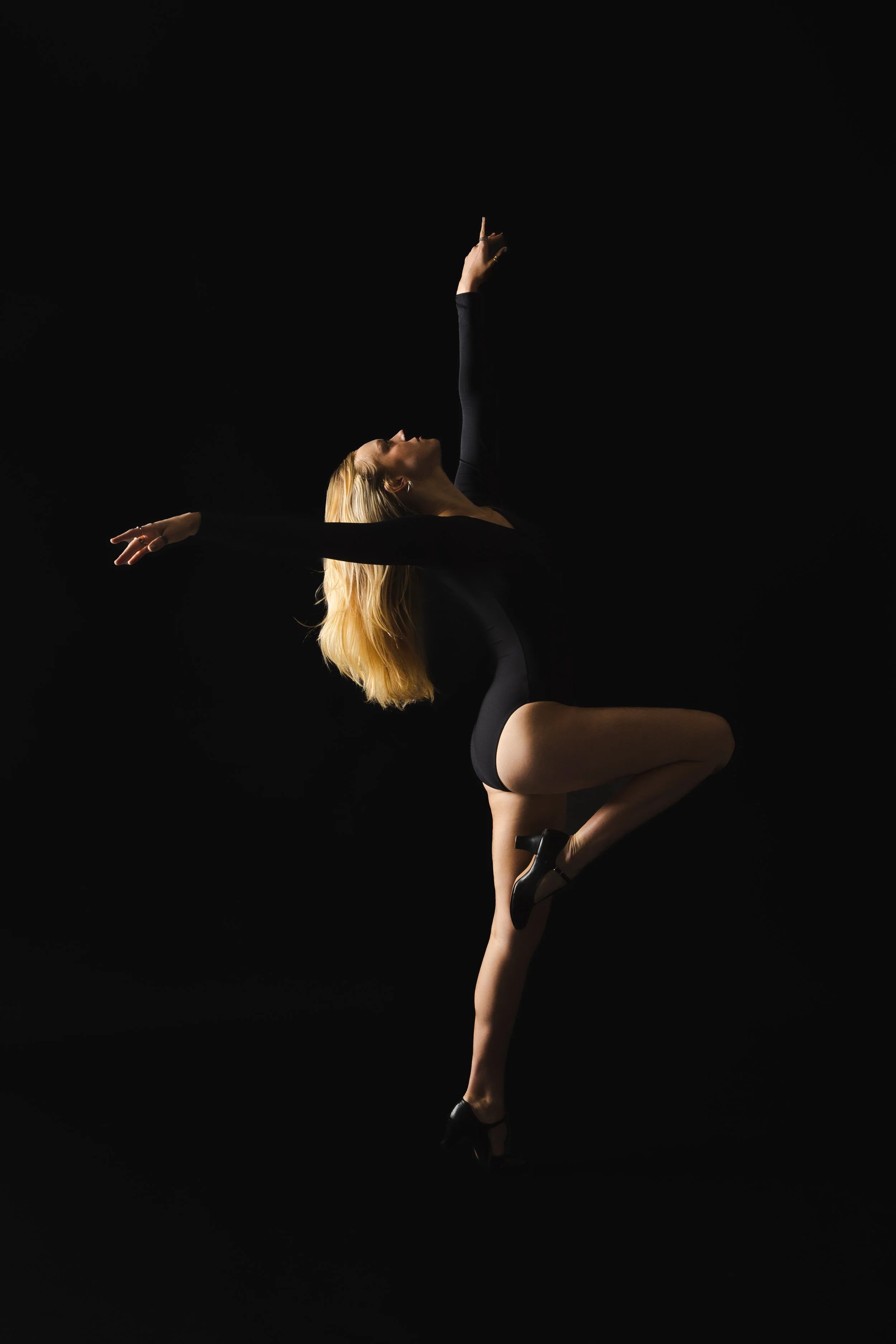 A woman with blonde hair performing a dance pose against a black background, wearing a black leotard and high heels.
