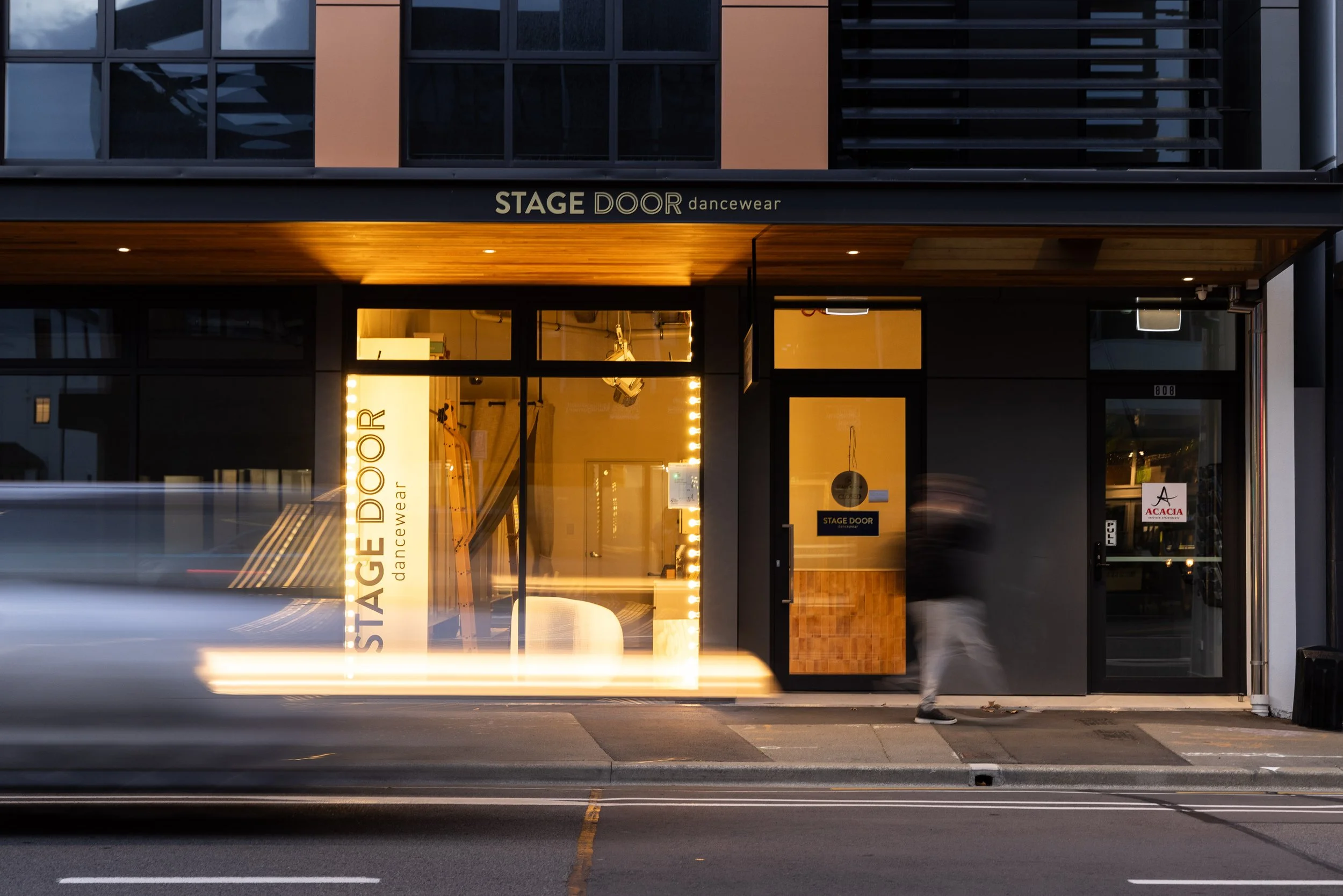 The storefront is for a dancewear shop called Stage Door. The shop has a modern black exterior with large glass windows showing inside lighting and dance equipment. There are street markings and a person walking past the shop.