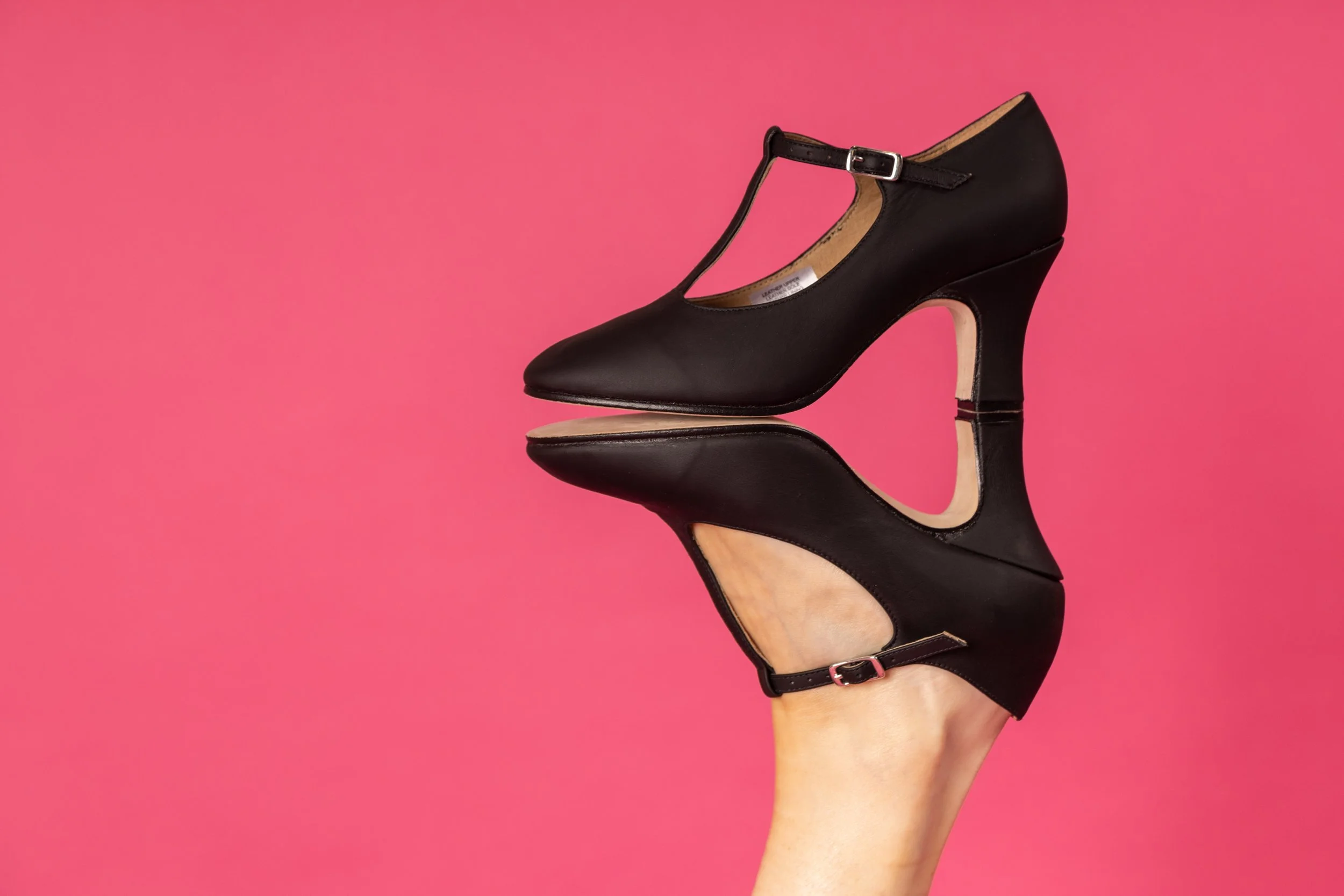 A person's hand raising a black high-heeled shoe with an ankle strap against a pink background.