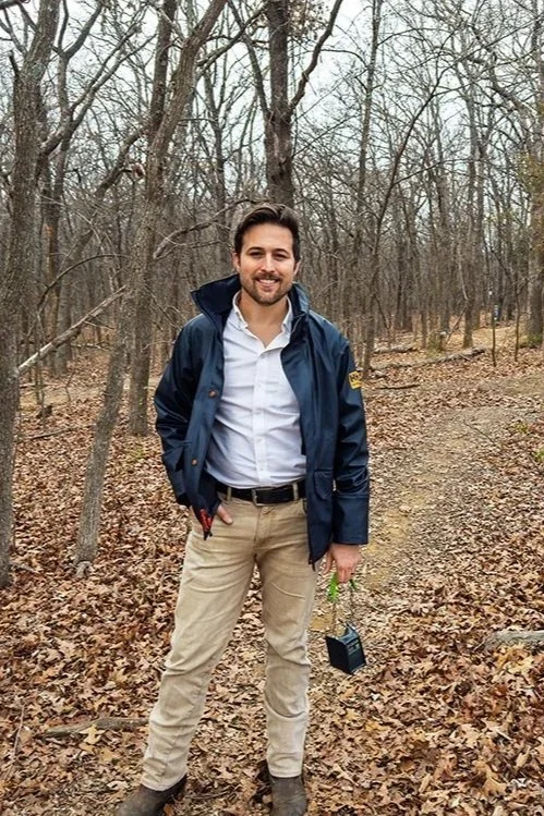 A man with dark hair and a beard, wearing a white shirt, beige pants, and a navy blue jacket, smiling and standing in a wooded area with leaf-covered ground.