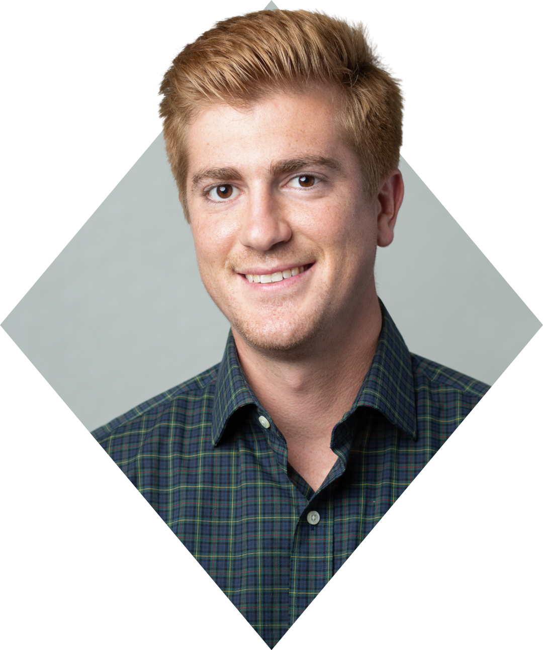 A young man with light skin, short blond hair, and a friendly smile, wearing a dark plaid shirt, against a plain gray background.