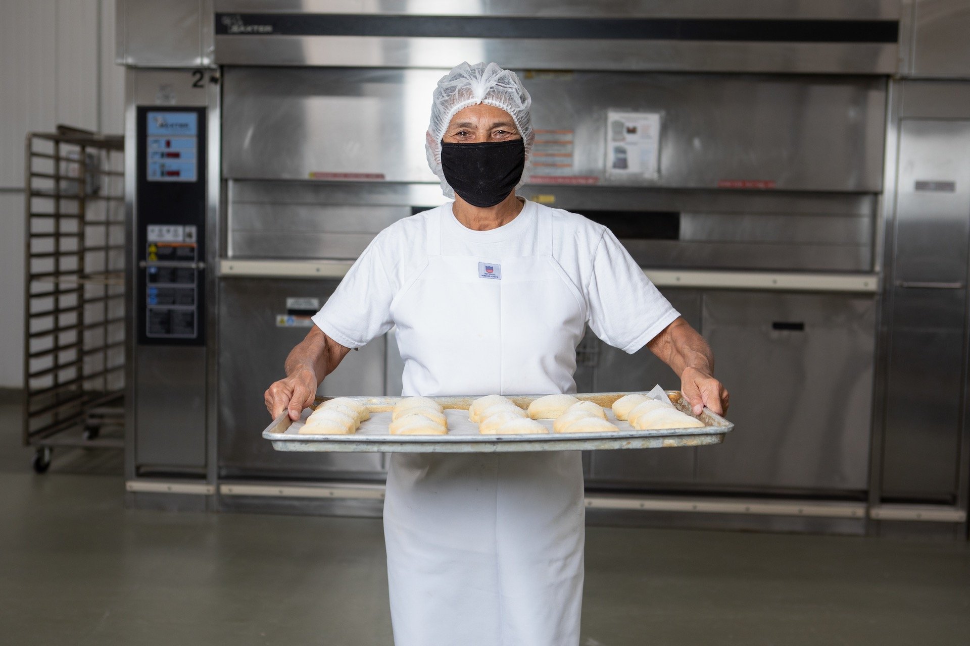 A woman in a hairnet and face mask holding a tray of raw bread dough in a commercial kitchen.