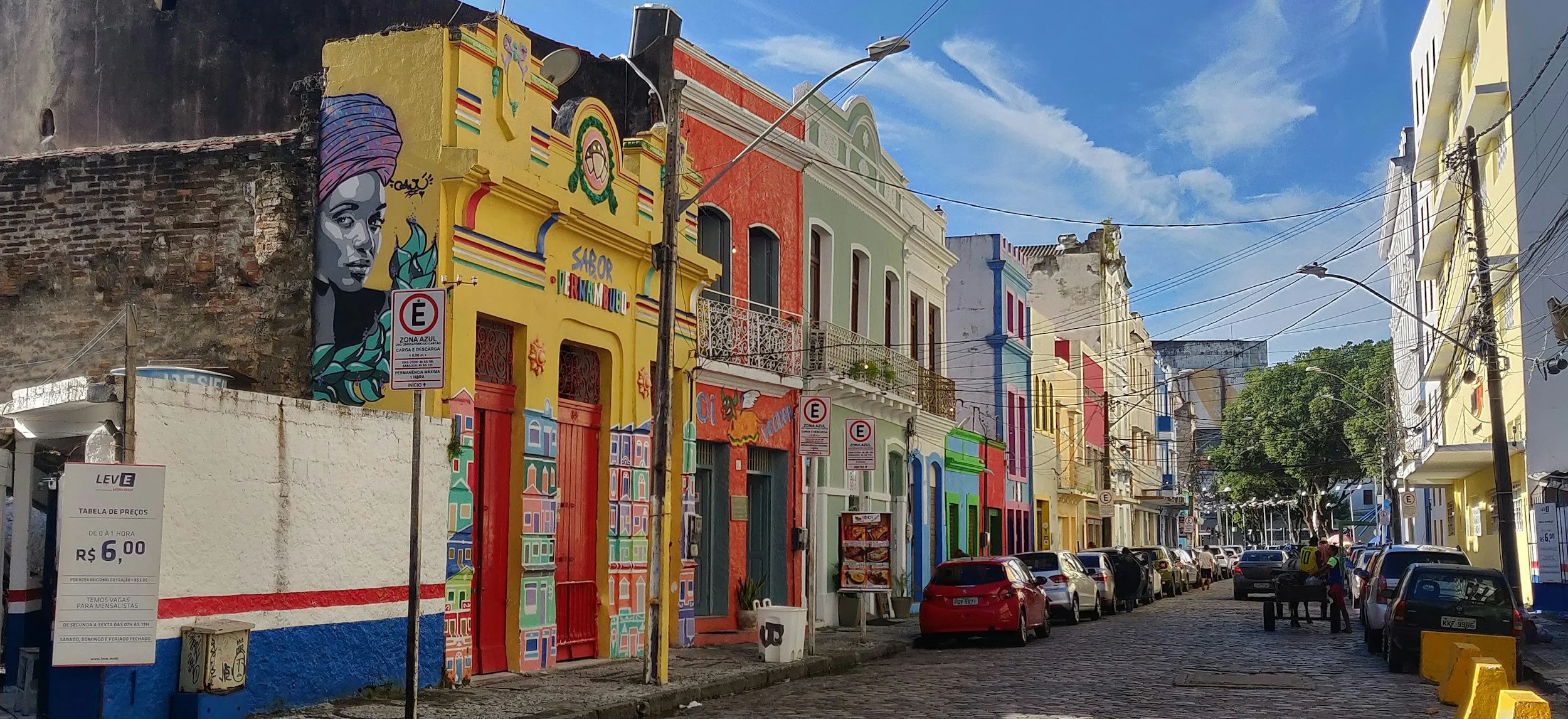 Colorful buildings with vibrant murals on a city street under a blue sky, with parked cars and some pedestrians.