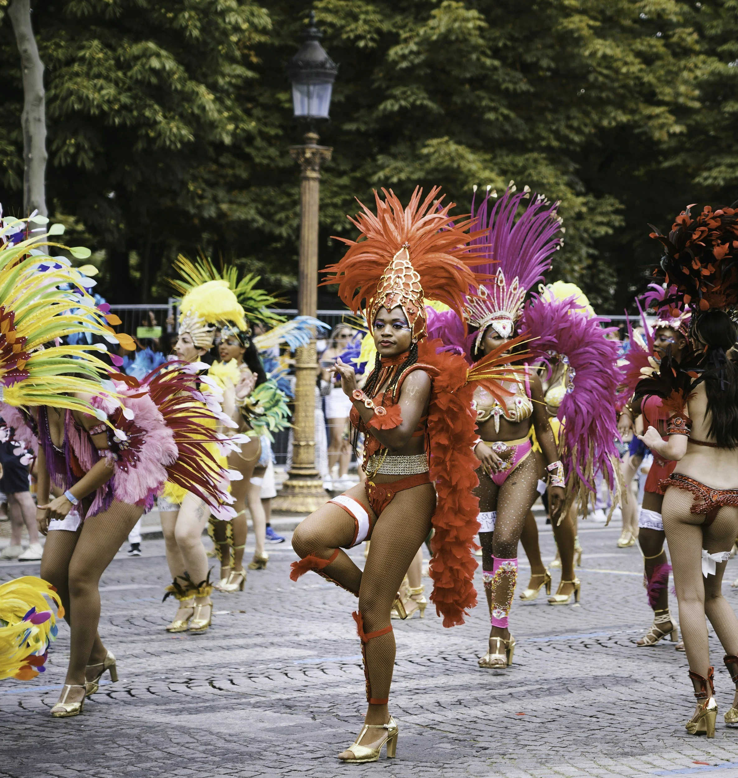 Women in colorful carnival costumes with feathered headdresses in a parade on a cobblestone street.