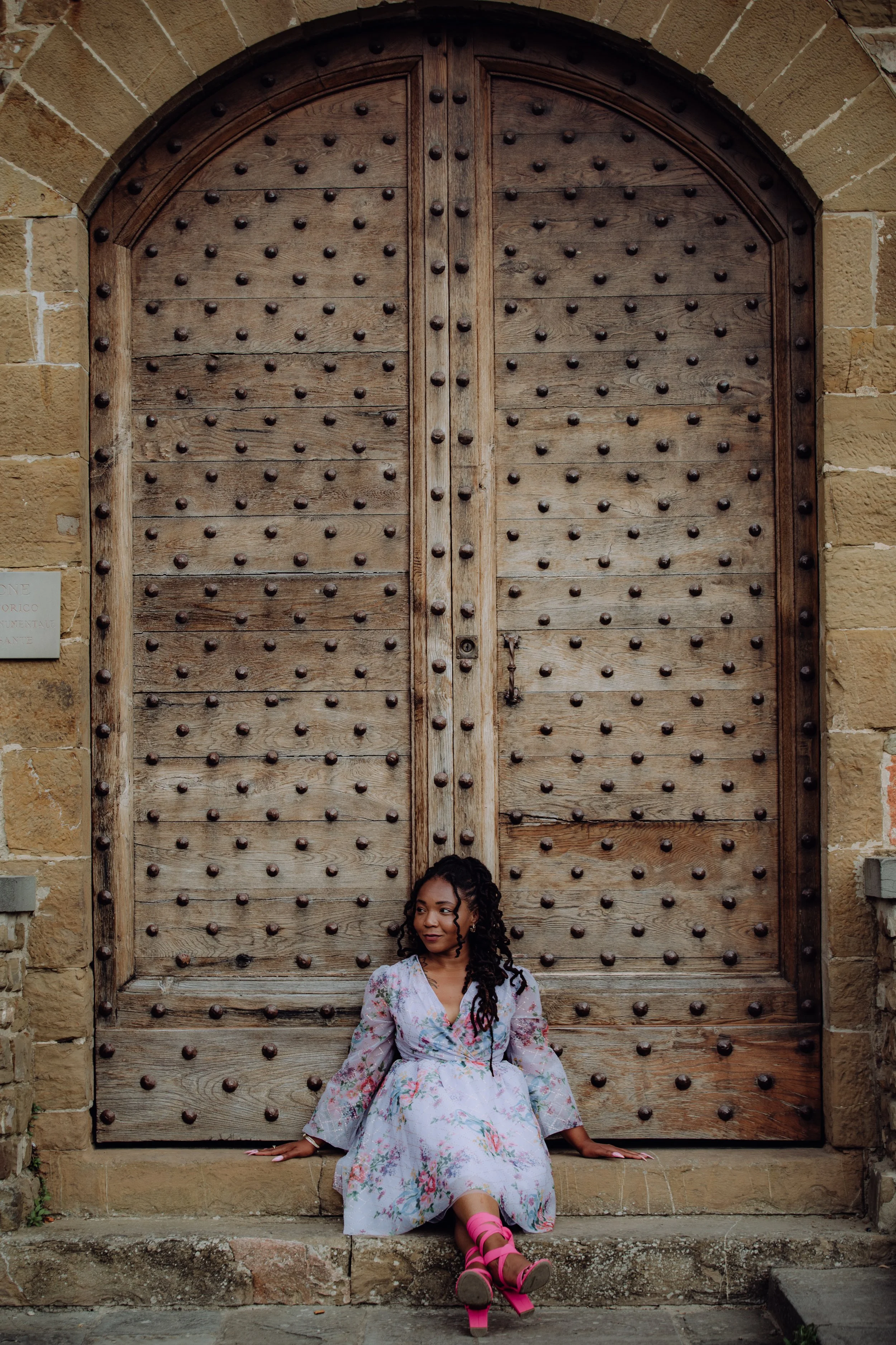 A woman with curly hair and floral dress sitting on stone steps in front of a large wooden door with metal studs, wearing pink high-heeled shoes.