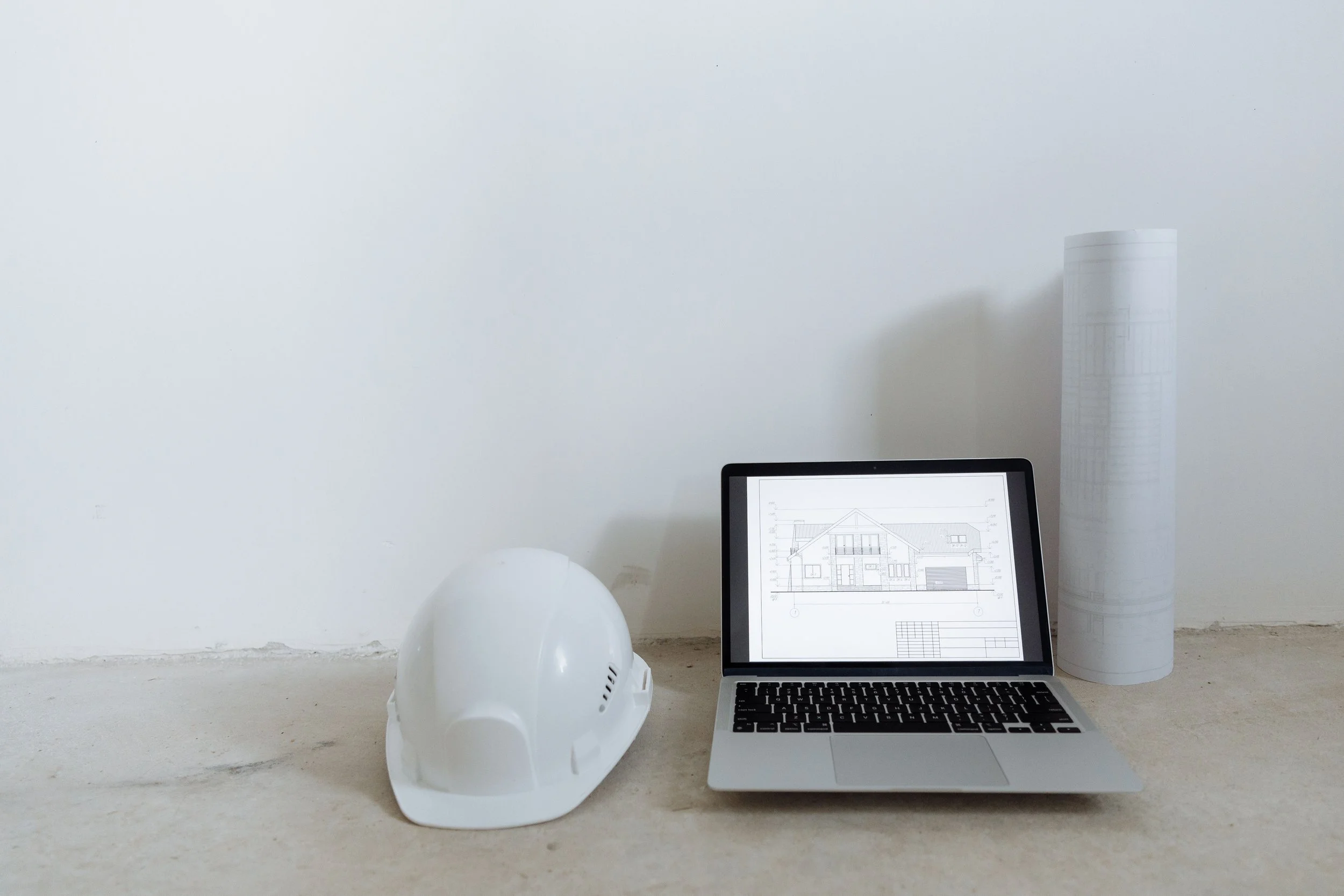 Casque de sécurité blanc, ordinateur portable avec plan d'architecture, rouleau de plans, et une bouteille d'eau, tout placé sur une surface en pierre dans un espace minimaliste.