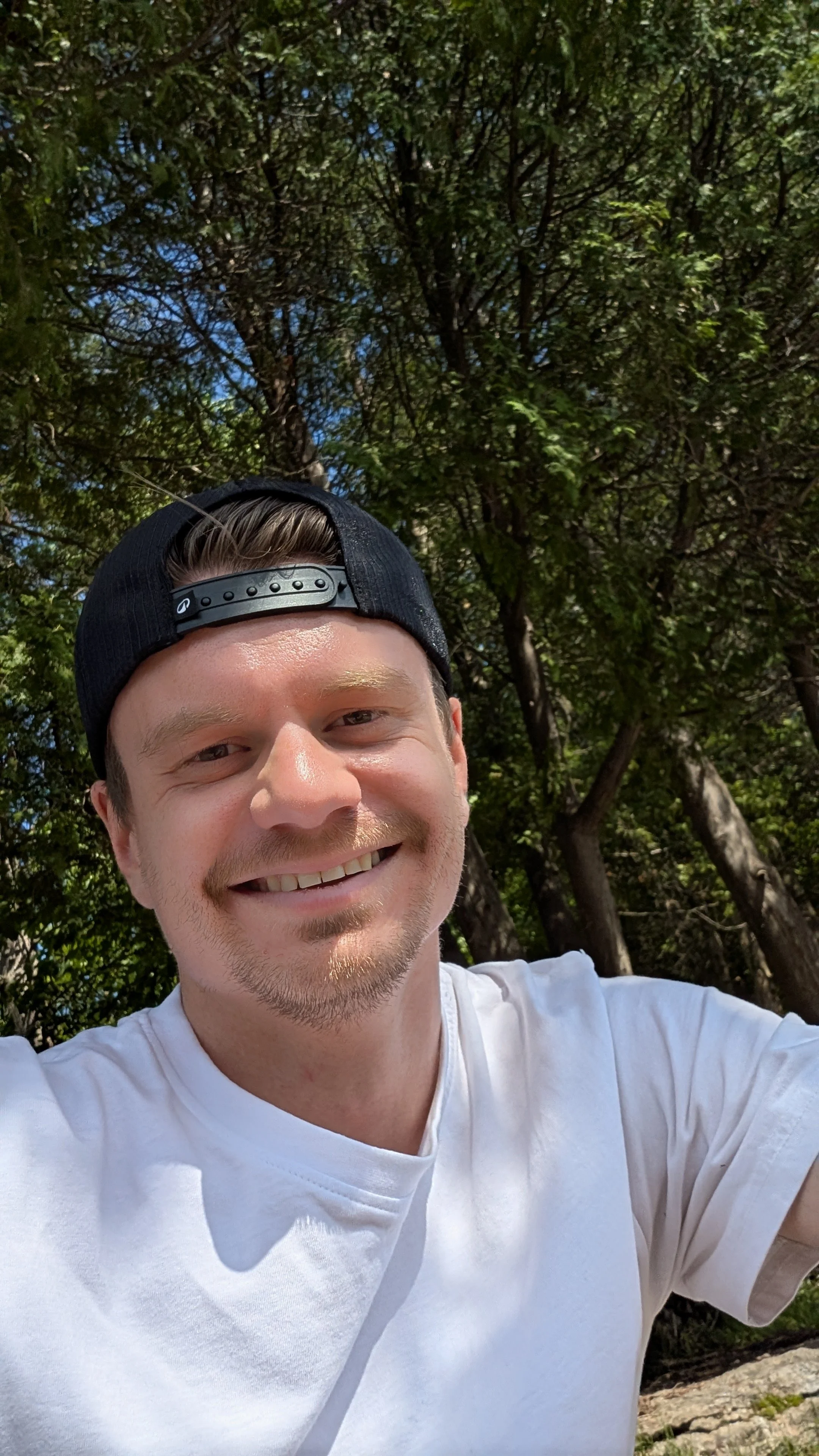 Jeune homme souriant en t-shirt blanc et casquette noire, prise en selfie dans un parc en plein air avec des arbres verts en arrière-plan, par temps ensoleillé.
