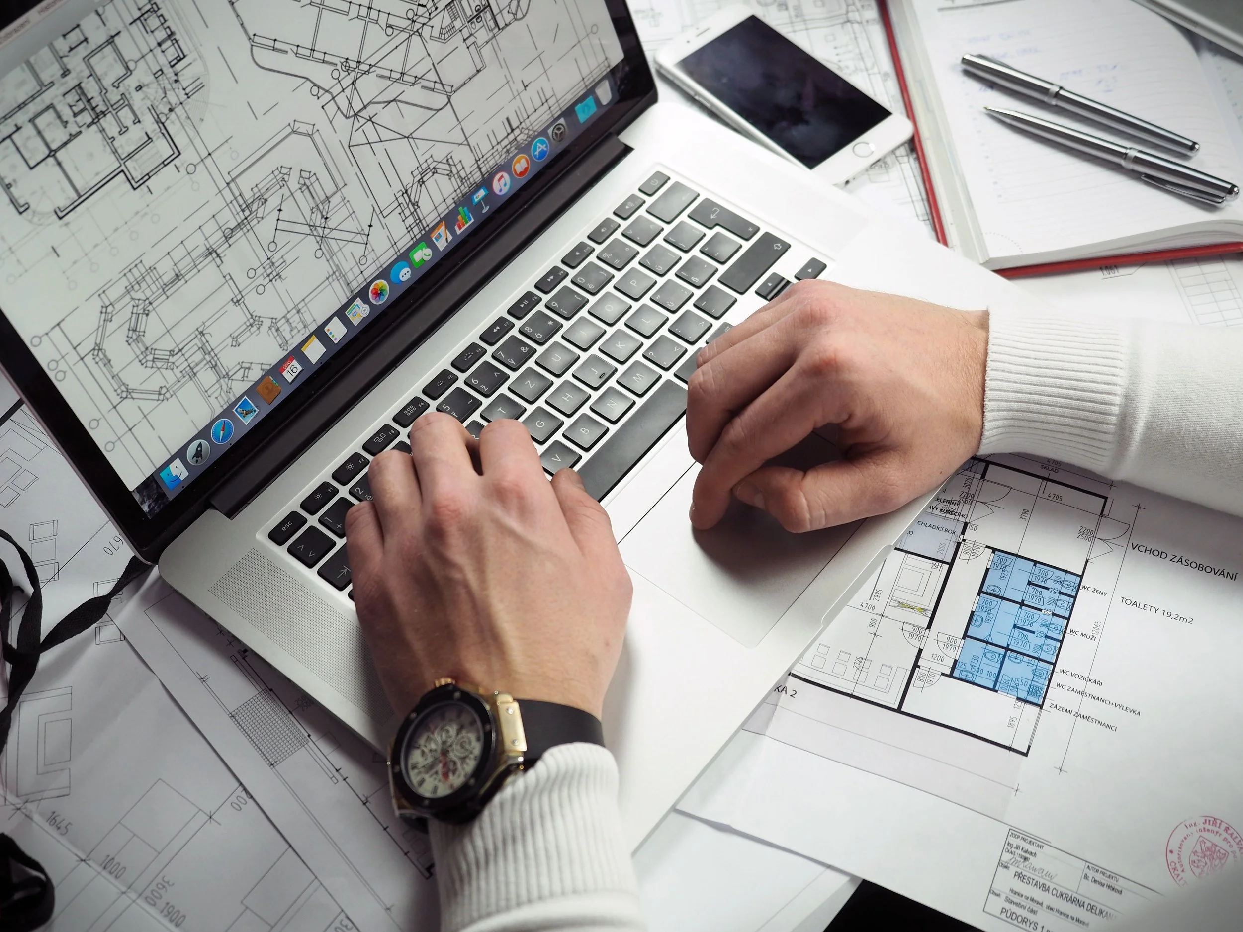 Une personne travaille sur un ordinateur portable avec des plans architecturaux étendus sous l'ordinateur, entourés de notes, stylos et un téléphone smartphone.