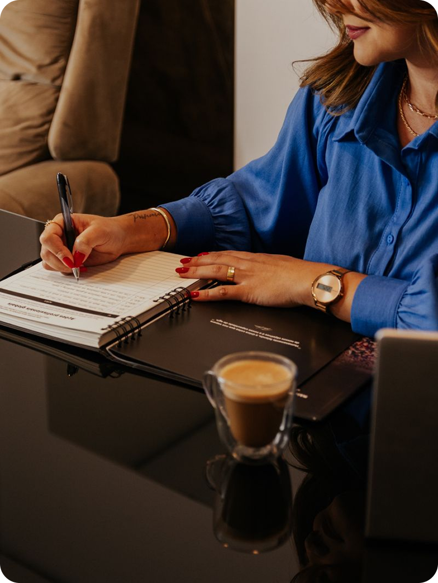 Woman in a blue shirt writing in a notebook with a pen, coffee on the table.