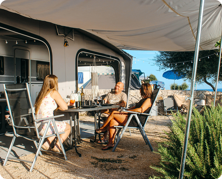 Three people sitting around a table outside of a camper, with drinks and food, under a large canopy, with a stone wall, trees, and blue sky in the background.