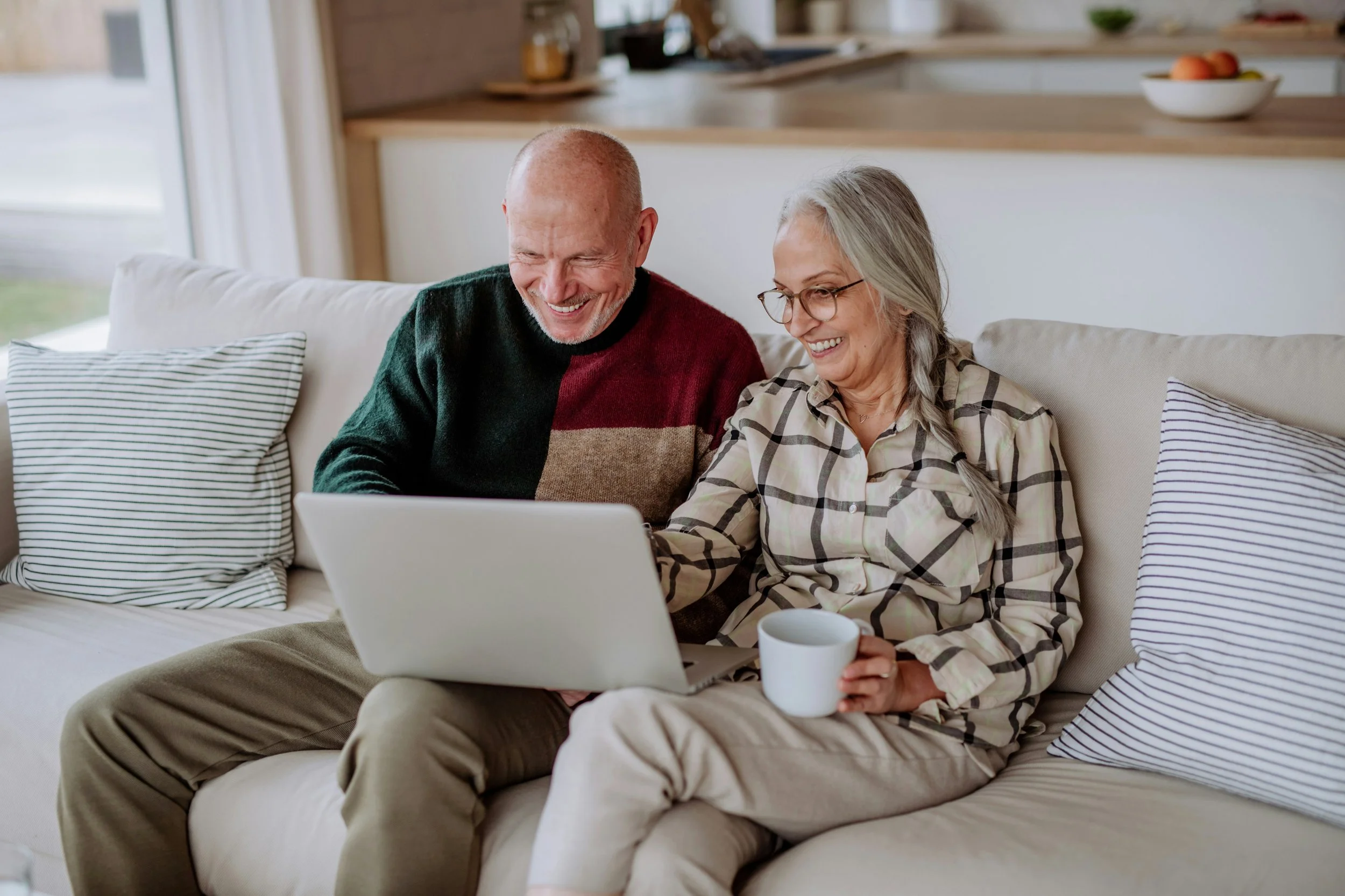 An elderly couple sitting on a beige sofa in a bright living room, smiling and looking at a laptop, with a mug in the woman's hand.
