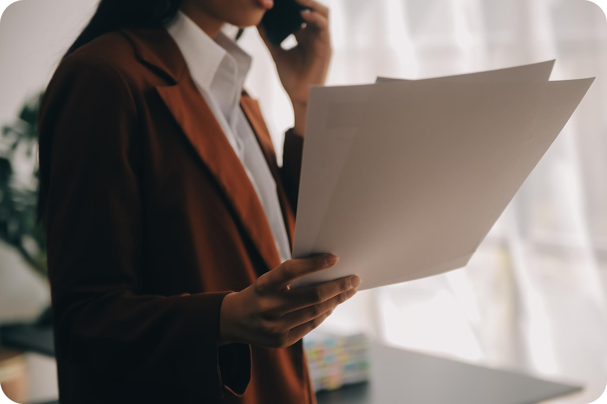 Woman in business attire reading a large document while on the phone in an office.