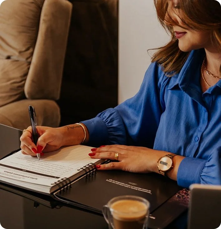 A woman with red hair, wearing a blue blouse, sitting at a desk and writing in a notebook with a black pen. She has red nail polish, a watch, and a ring, and there is a cup of coffee on the desk.