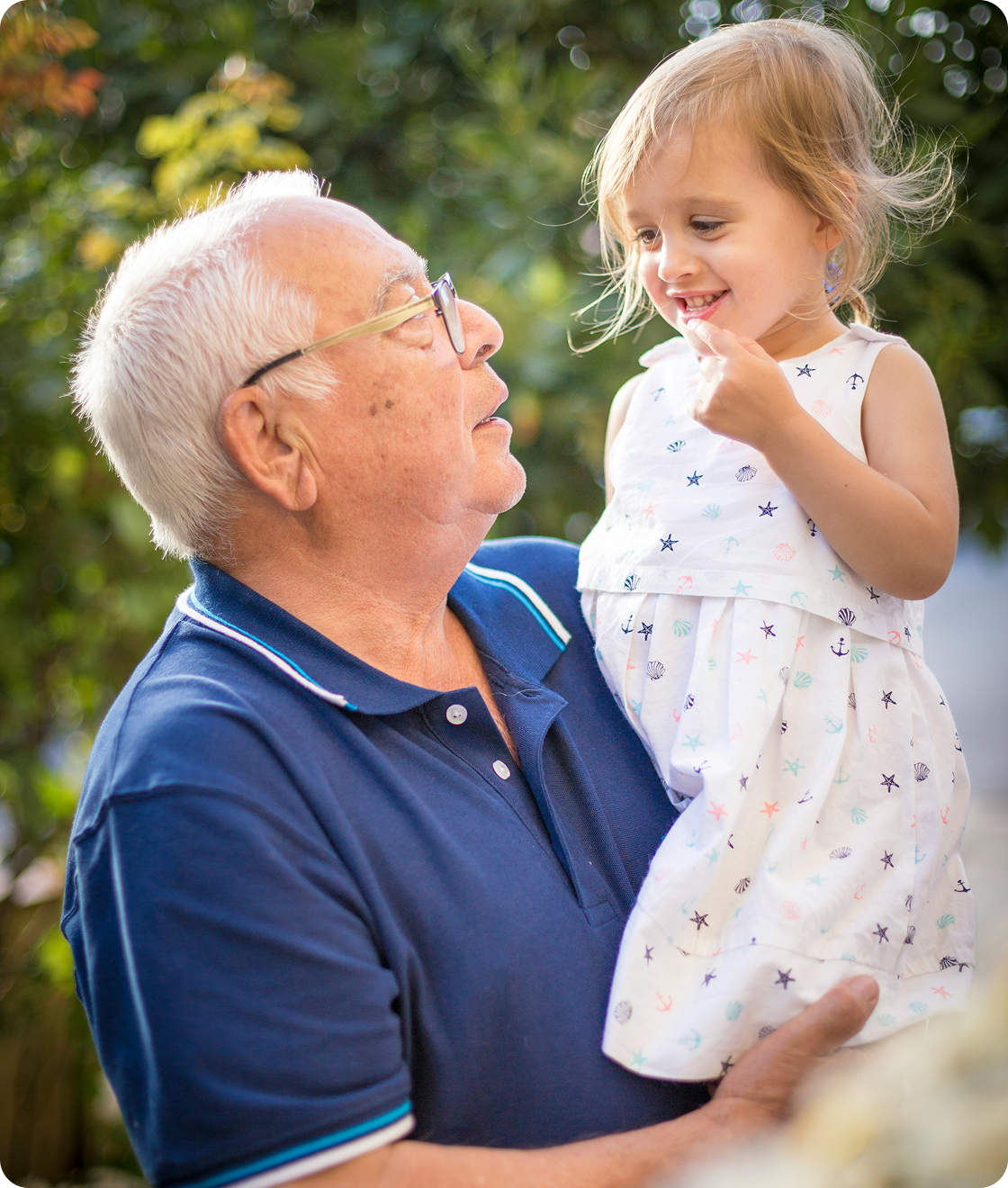 An elderly man with glasses holding a young girl in his arms, outdoors with greenery in the background.
