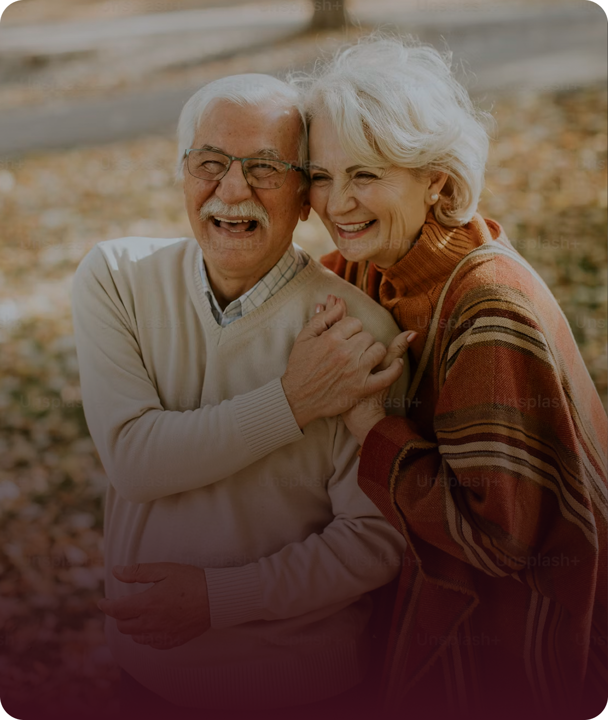 An elderly couple smiling and laughing together outdoors, embracing each other, with autumn leaves on the ground.