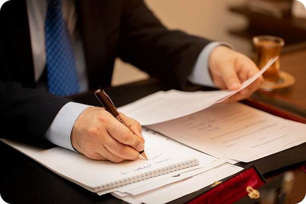 A person in a suit and blue tie is writing on a notepad with a pen, holding a paper with the other hand, on a desk with documents and a tea cup.
