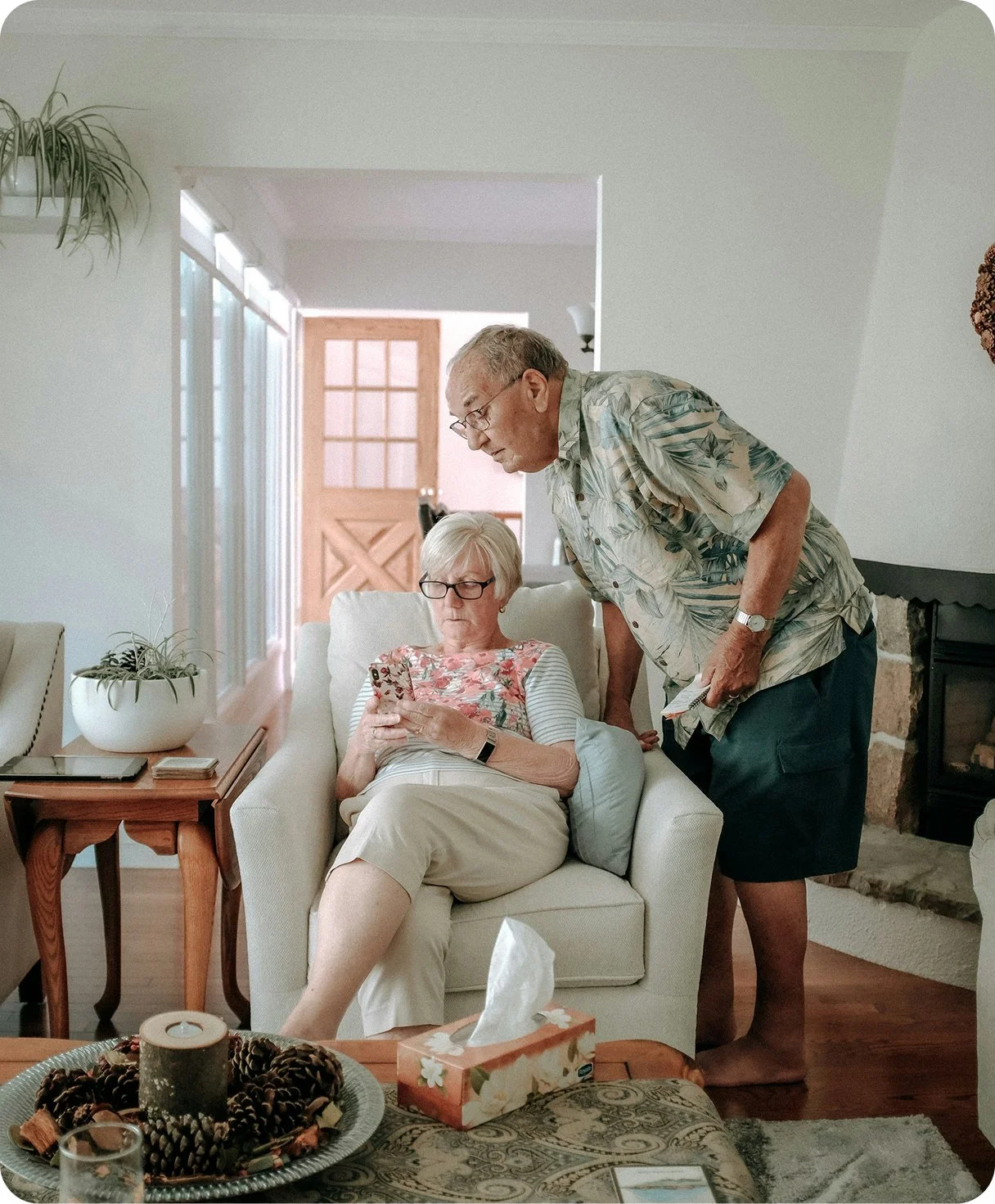 An elderly woman sitting on a beige armchair looking at her cell phone, while an elderly man stands beside her leaning over to look at her phone. They are in a well-lit living room with windows, a side table with a plant, and a fireplace.